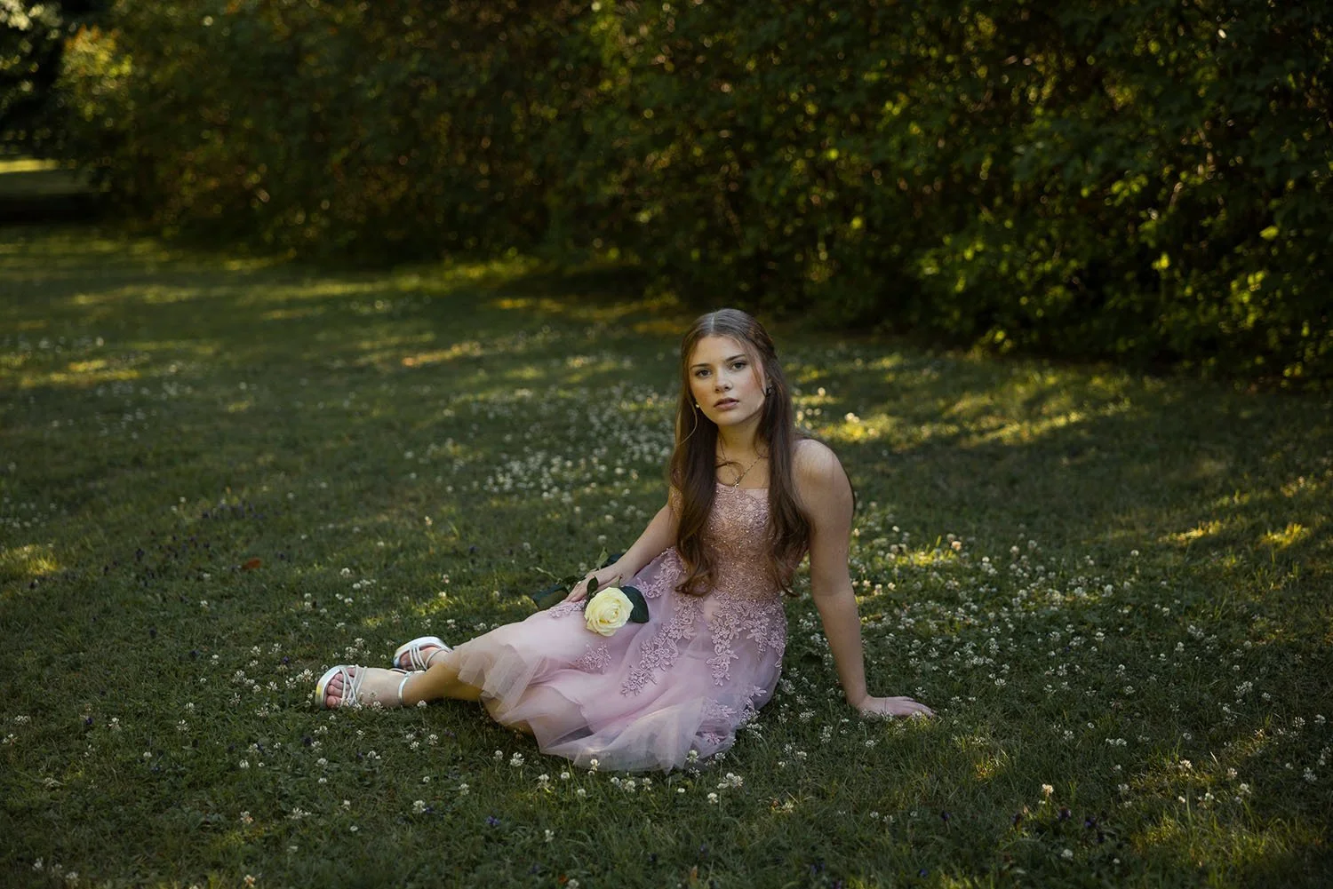 Tunnelmallinen rippikuvaus luonnossa – nuori nainen istuu ruohikolla vaaleassa juhlamekossa.