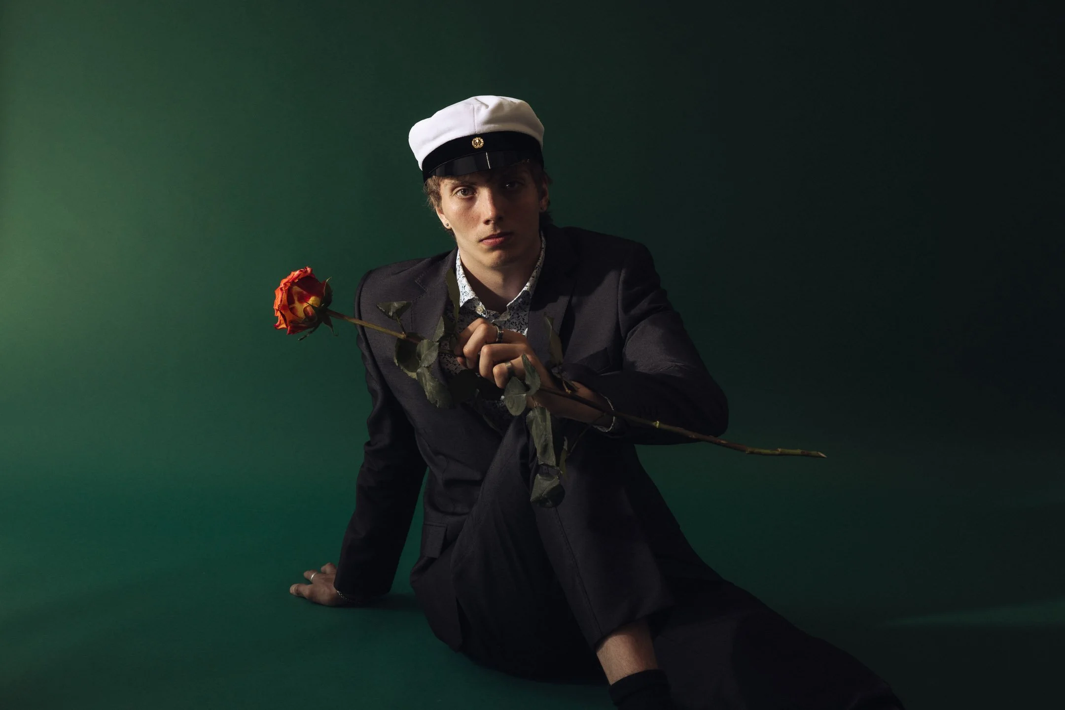 Tyylikäs valmistujaiskuvaus studiossa – ylioppilasasussa oleva nuori mies istuu ruusun kanssa vihreällä taustalla.