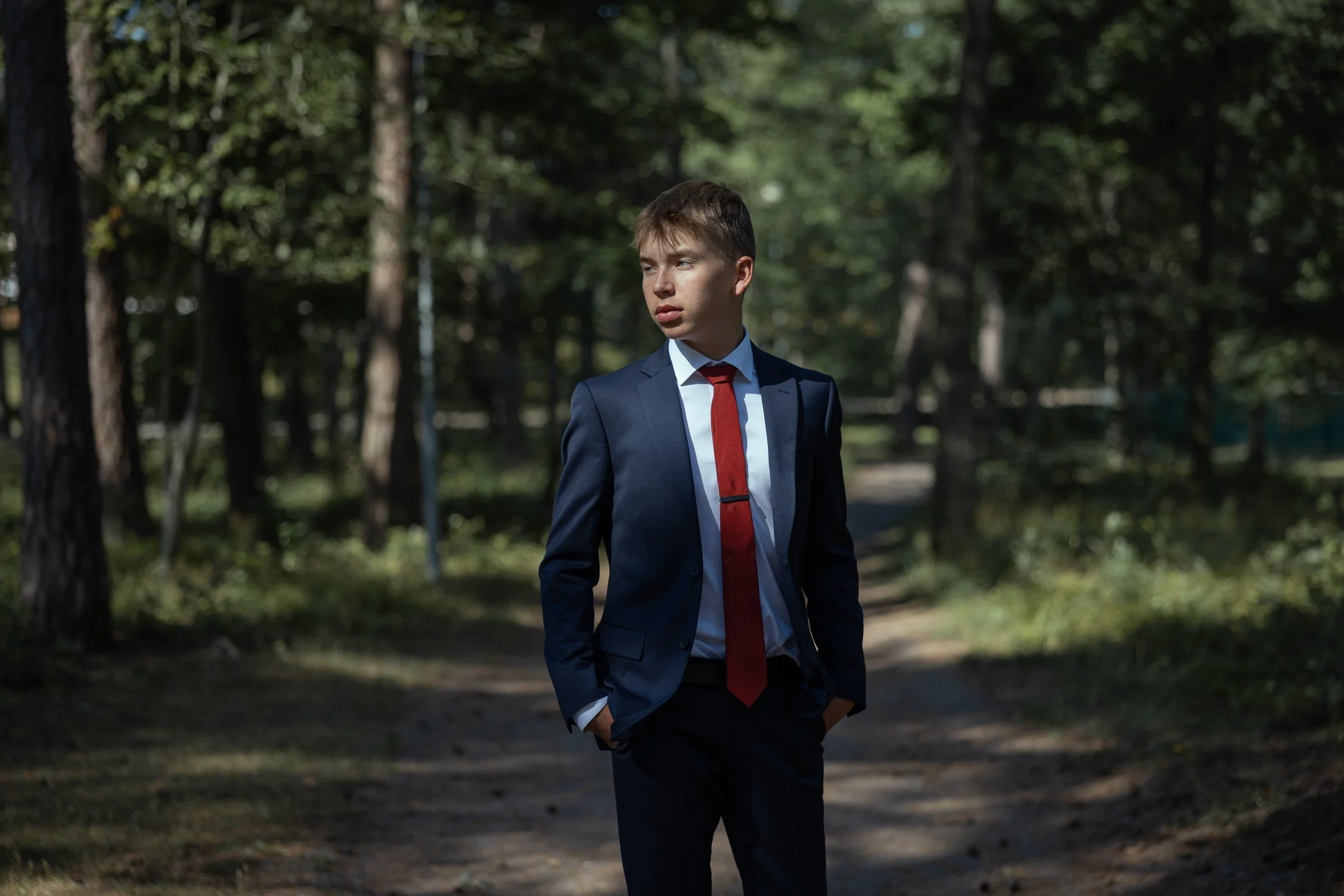 Ammattimainen rippikuvaus ulkona – nuori mies puvussa ja punaisessa kravatissa metsäisessä miljöössä.