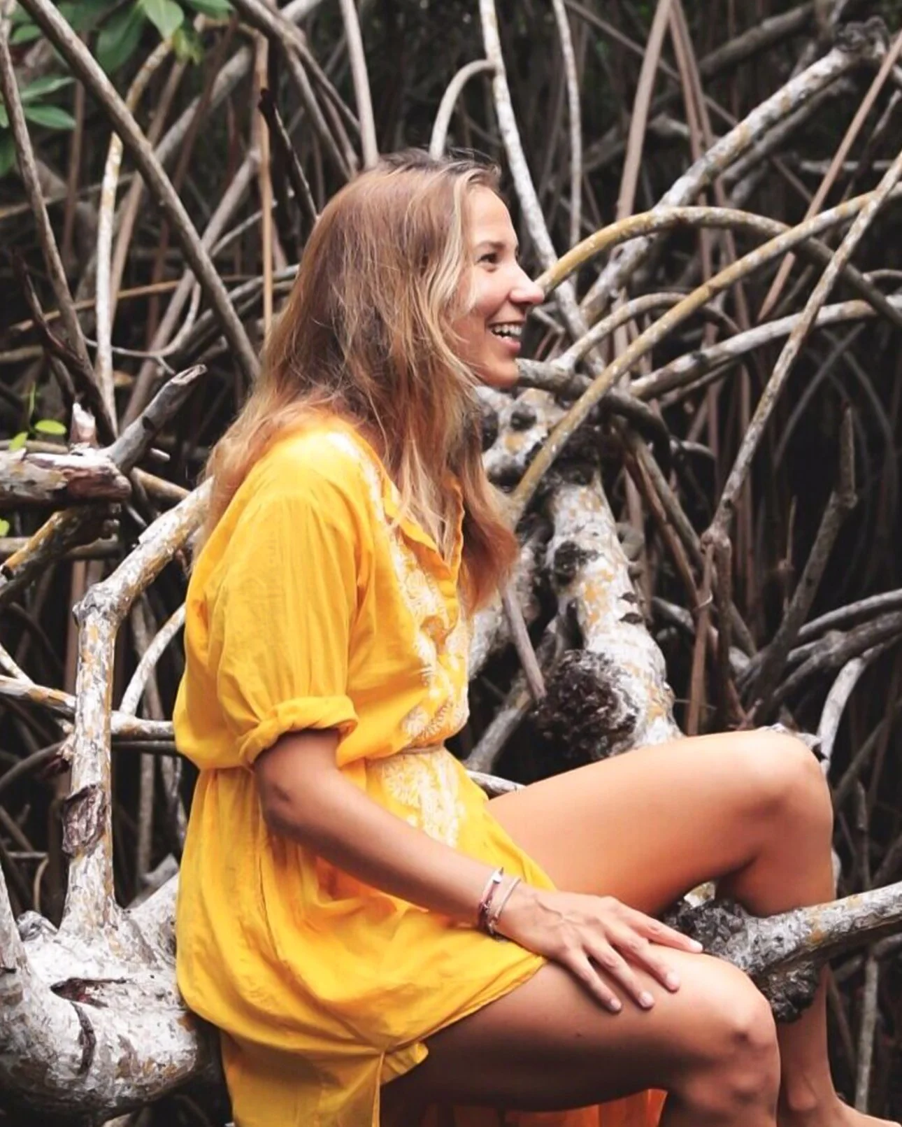 A woman wearing a yellow dress sitting on a fallen tree trunk in a mangrove forest, smiling and looking to her left.