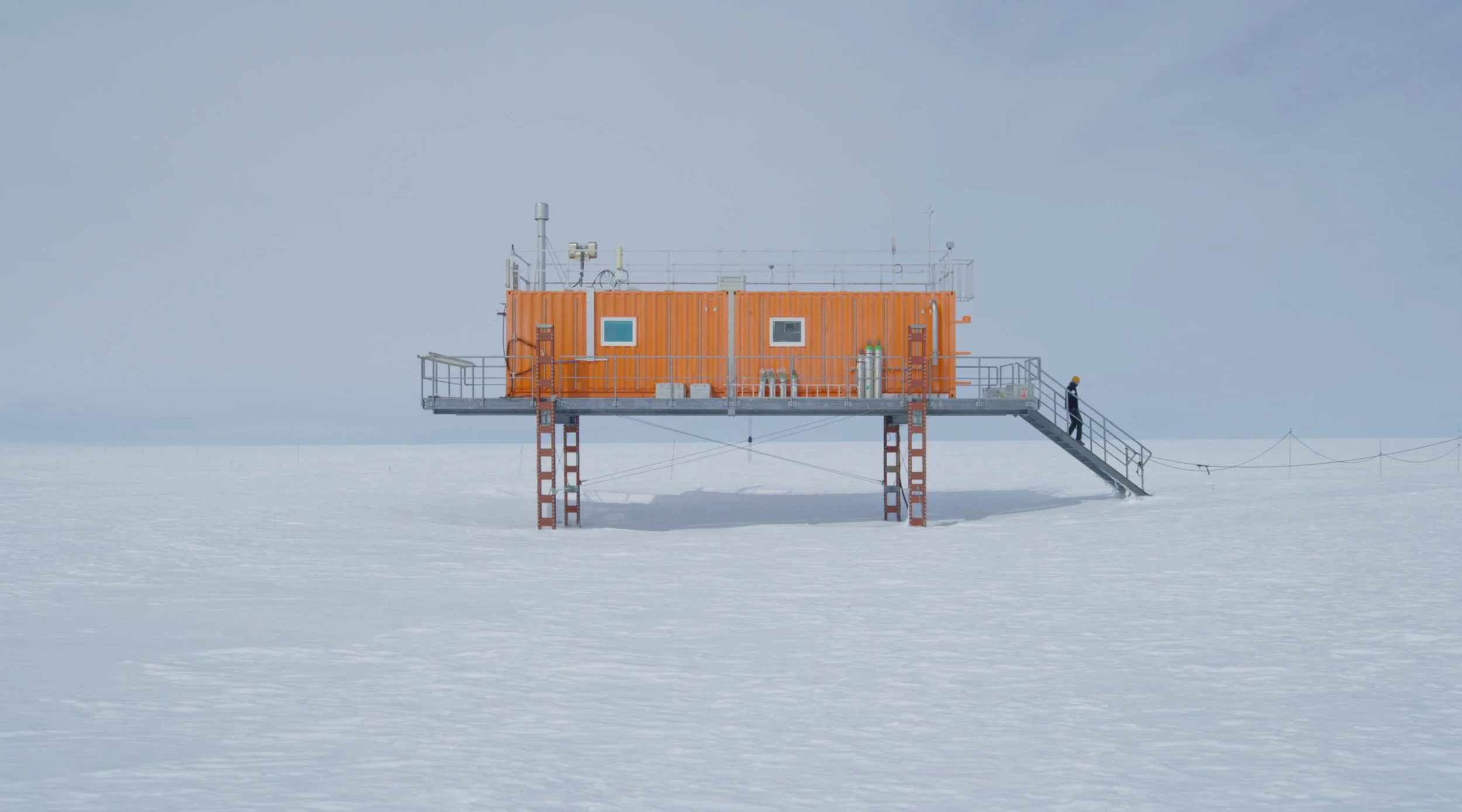 A scientific research station on stilts in a snowy, icy landscape, with orange container-like structures and a person walking up the stairs.