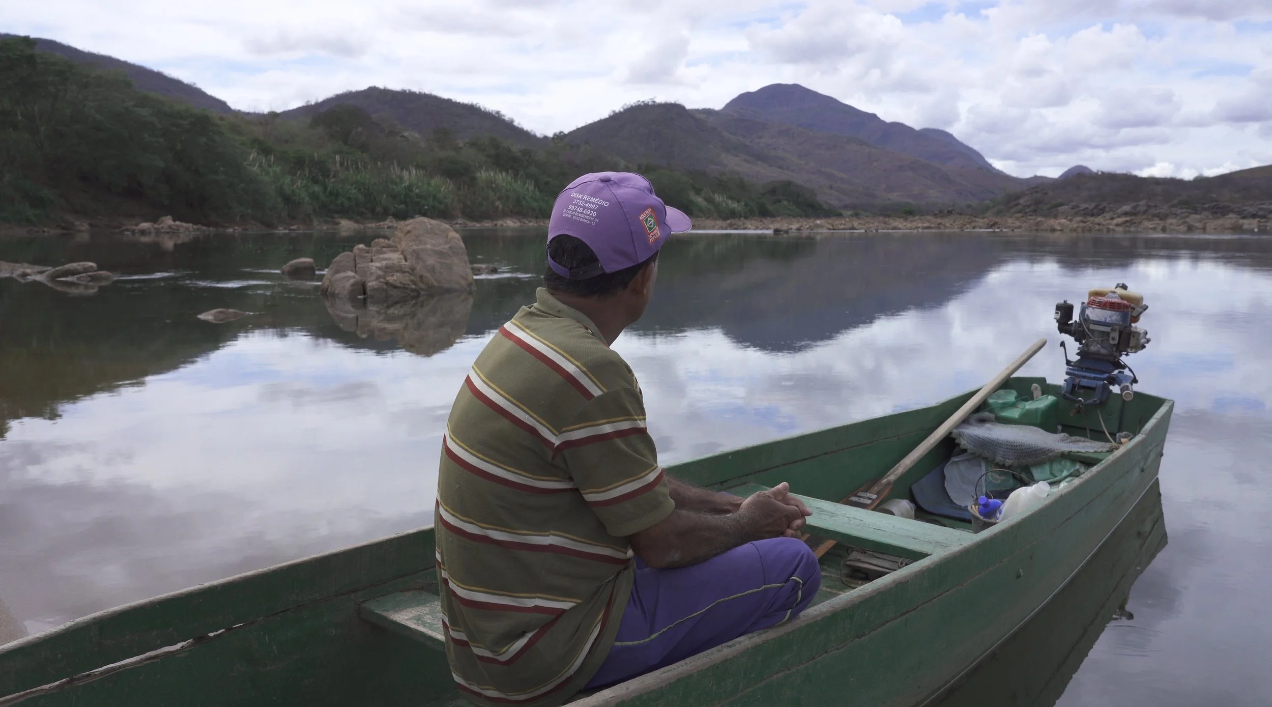 A man sitting in a green boat with a motor, on a calm lake with reflections of the cloudy sky and surrounding mountains, wearing a purple cap and a striped shirt.