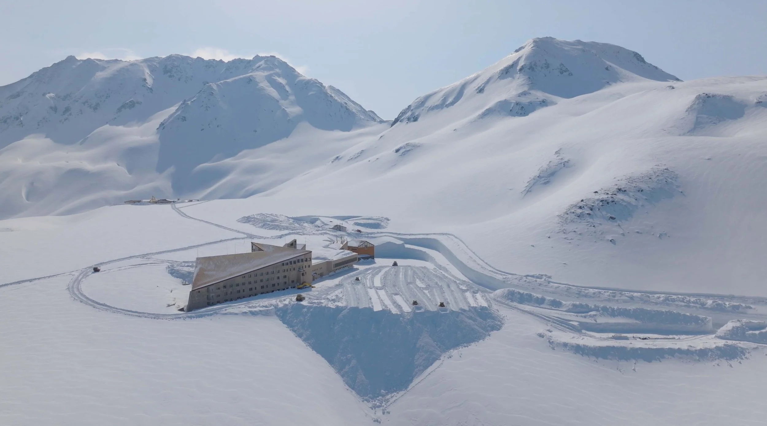 A snow-covered mountain landscape with a large building and multiple smaller structures, with ski tracks and snow vehicles in the foreground.