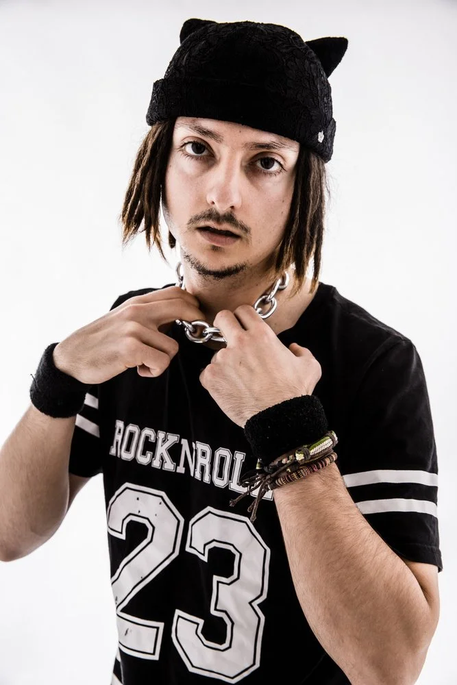 Young man with shoulder-length brown hair, wearing a black beanie with small cat ears, a black T-shirt with white text and numbers, multiple bracelets, and a silver chain necklace. He has a slightly parted mouth and is looking directly at the camera against a plain white background.