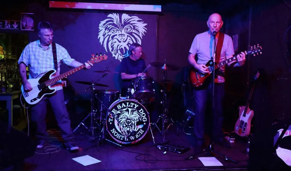 Three musicians performing on stage in a dimly lit venue with a lion logo on the backdrop and a drum set that says "The Salty Dog".