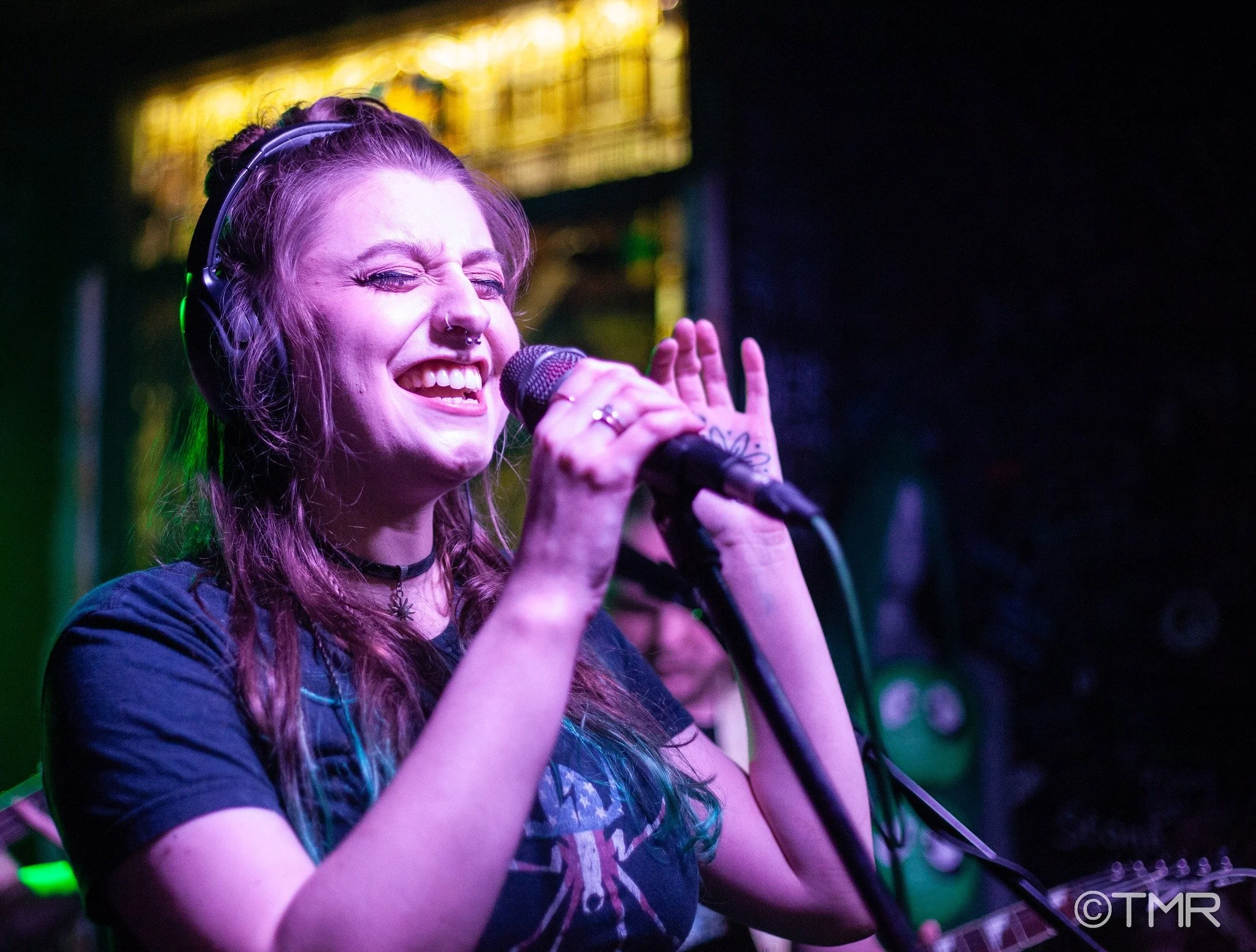 A female singer wearing headphones, smiling and singing into a microphone on stage with colorful lights in the background.