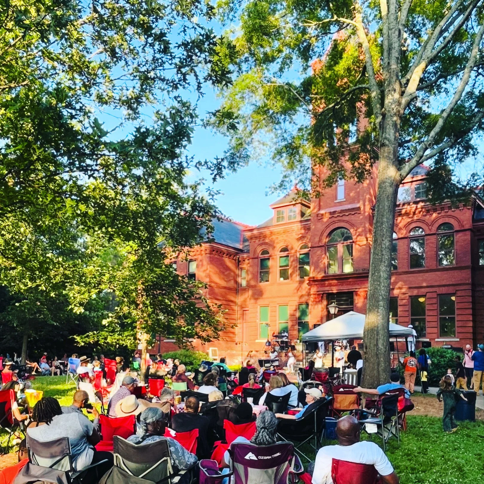 A gathering of people sitting on chairs and blankets on a grassy area in front of a red brick building, with trees and a clear blue sky in the background.