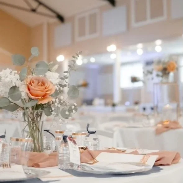 A table setup for a formal event with a floral centerpiece, plates, glasses, and napkins in a bright, decorated hall.