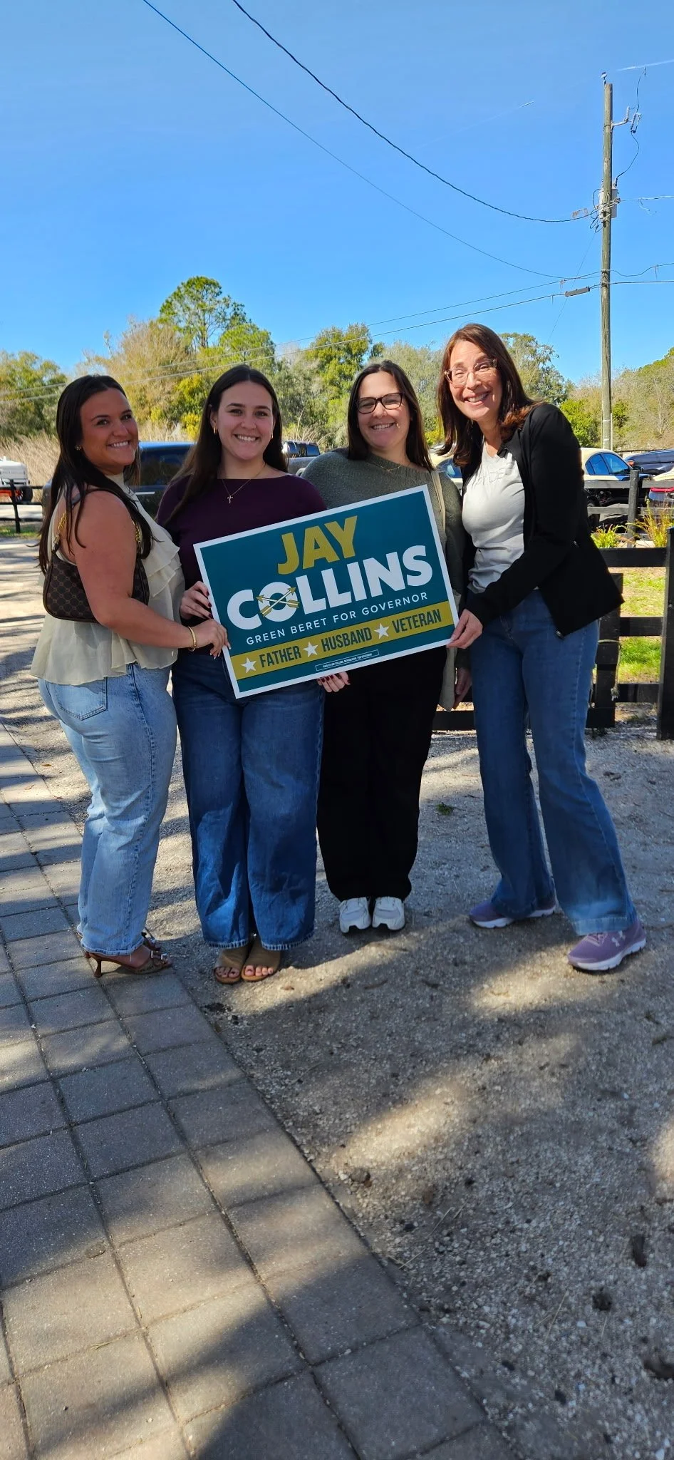 Elissa and three other women standing outdoors on a sunny day, holding a campaign sign that reads "Jay Collins, Green Beret for Governor, Father, Husband, Veteran." They are smiling and dressed casually, with trees and parked cars in the background.
