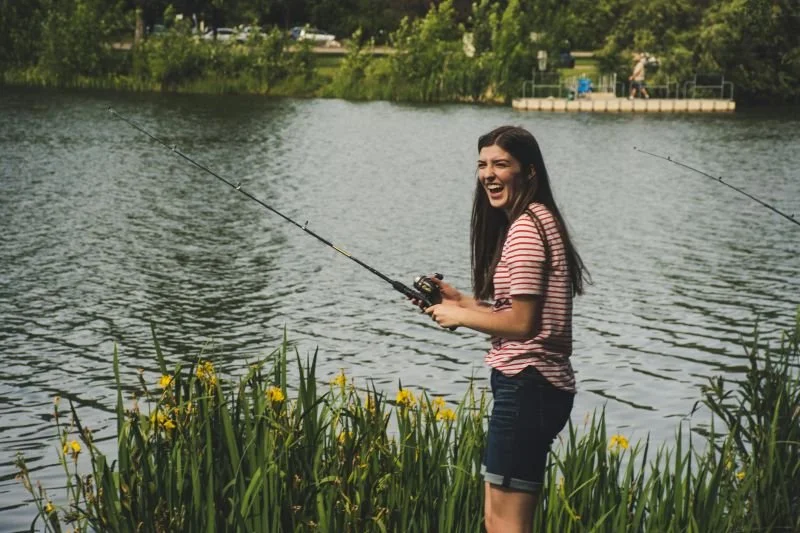 A young woman standing by a lake, holding a fishing rod and smiling.