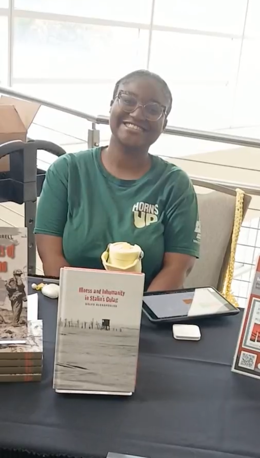 A woman, USF volunteer, sitting at the sign-in table, smiling, wearing glasses and a green t-shirt that says 'Horns Up', with books and promotional materials displayed on the table.