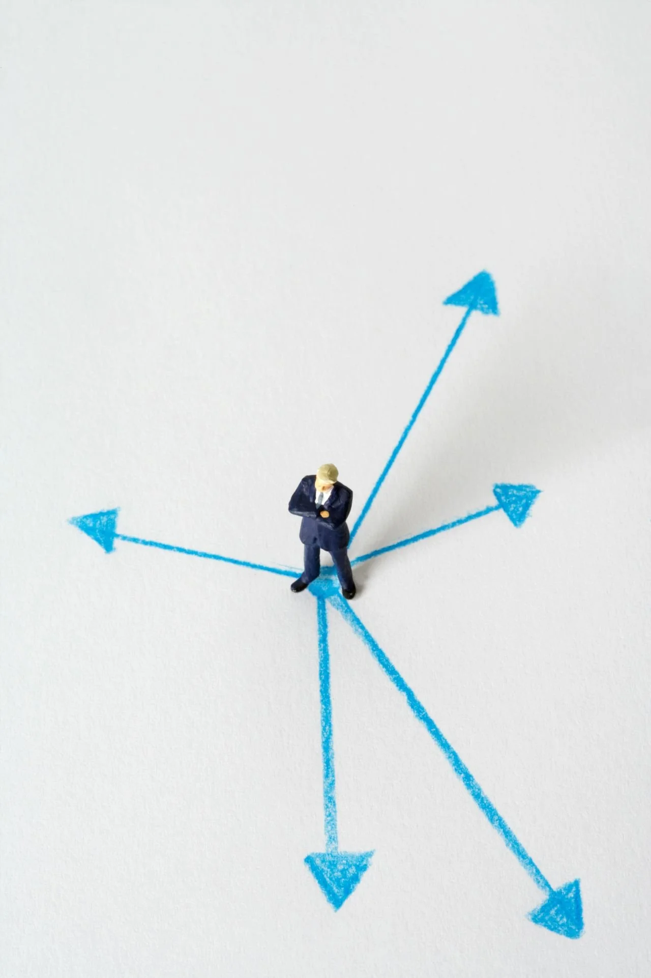 Miniature man in business suit standing on a blue arrow, with more blue arrows pointing in different directions on a white background.