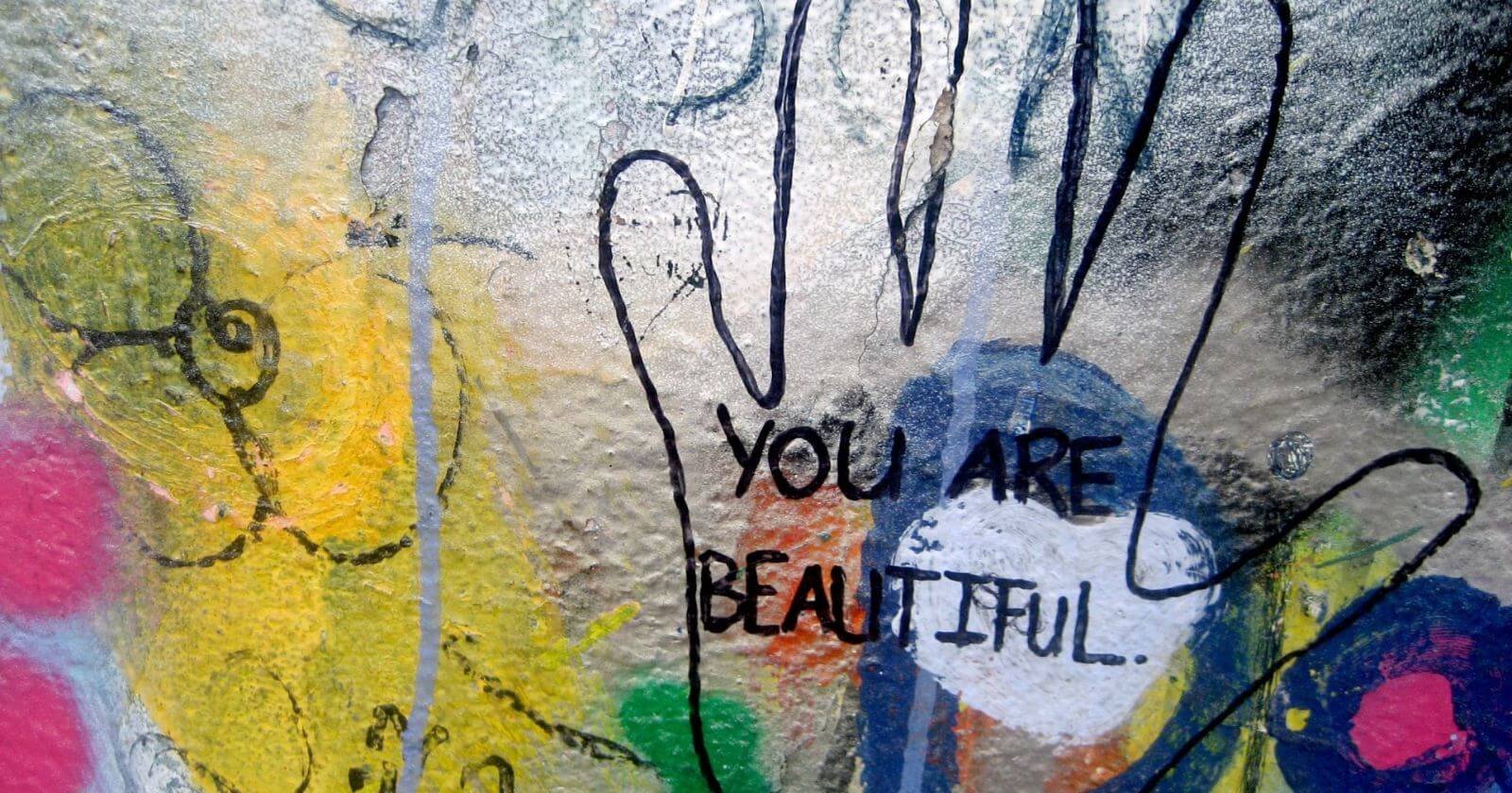 Outline of a hand against frosted, graffitied glass with the inspirational words You Are Beautiful printed inside.