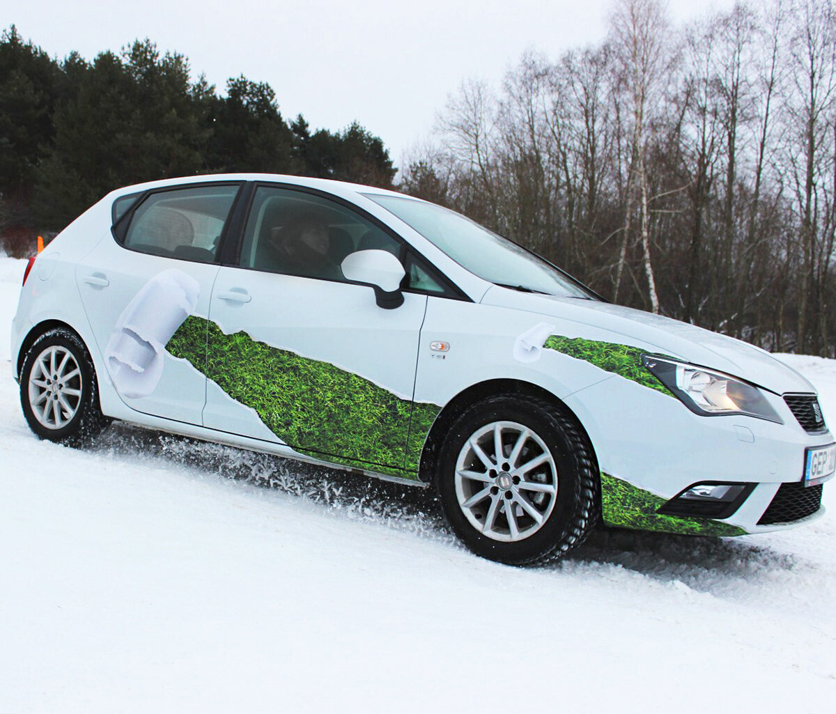 White car with green moss pattern and white foam design parked on snowy ground, with a background of leafless trees and an overcast sky.