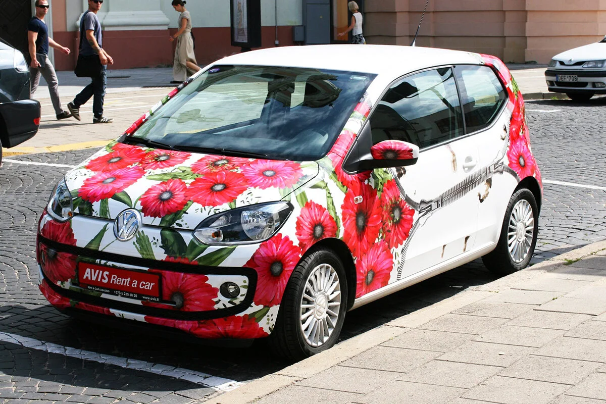 Small Volkswagen car covered in a vibrant floral wrap with red and pink flowers and green leaves, parked on a city street with pedestrians walking in the background.