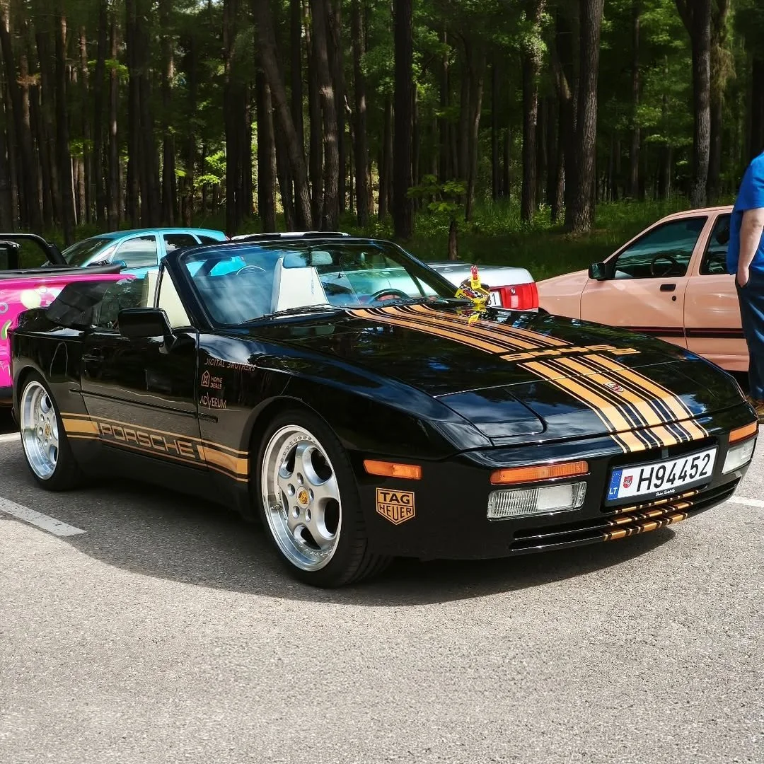 A black Porsche convertible with gold racing stripes parked in a lot with other cars, set against a background of trees.