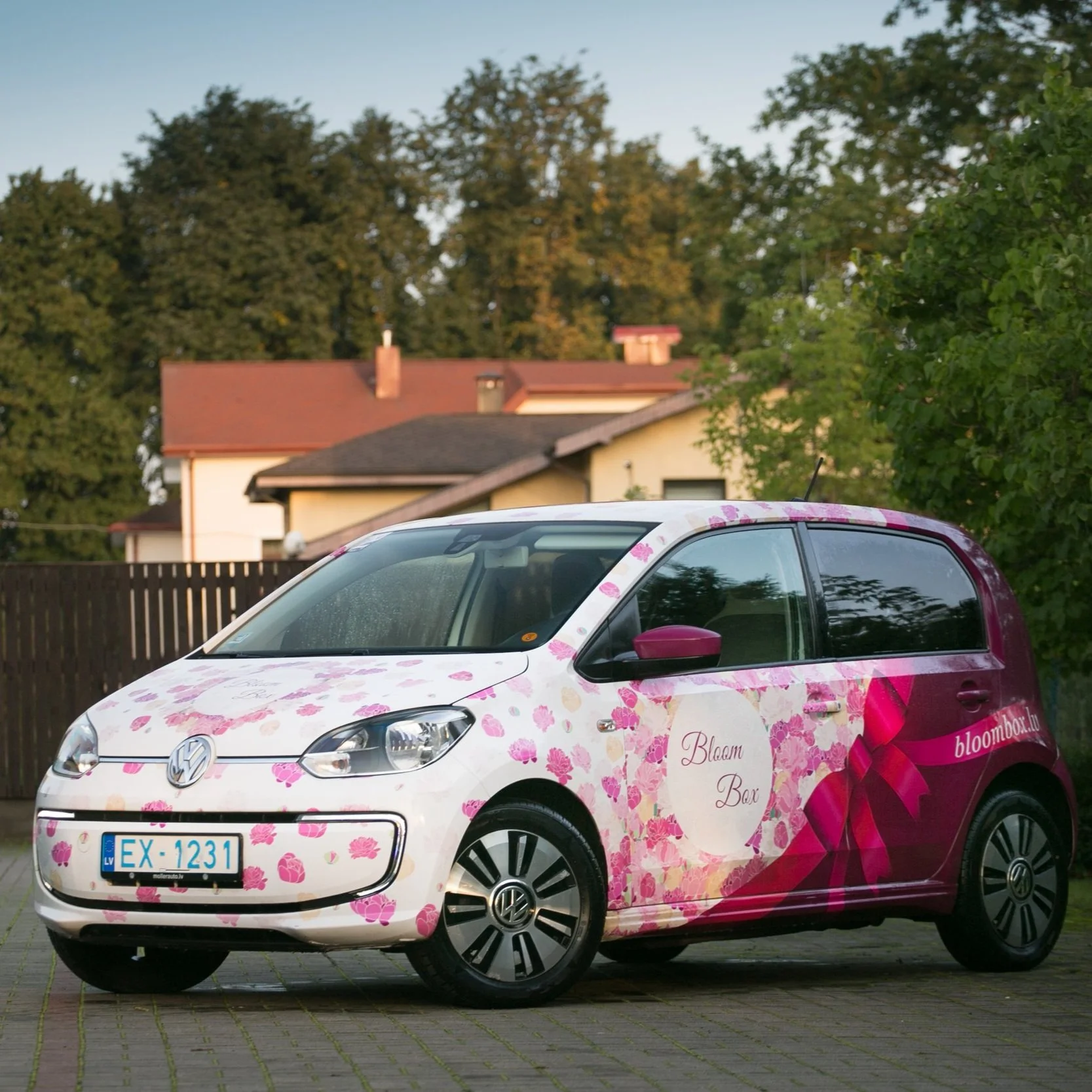 A Volkswagen car decorated with pink floral patterns and promotional branding for Bloom Box, parked outdoors on a paved surface with trees and houses in the background.