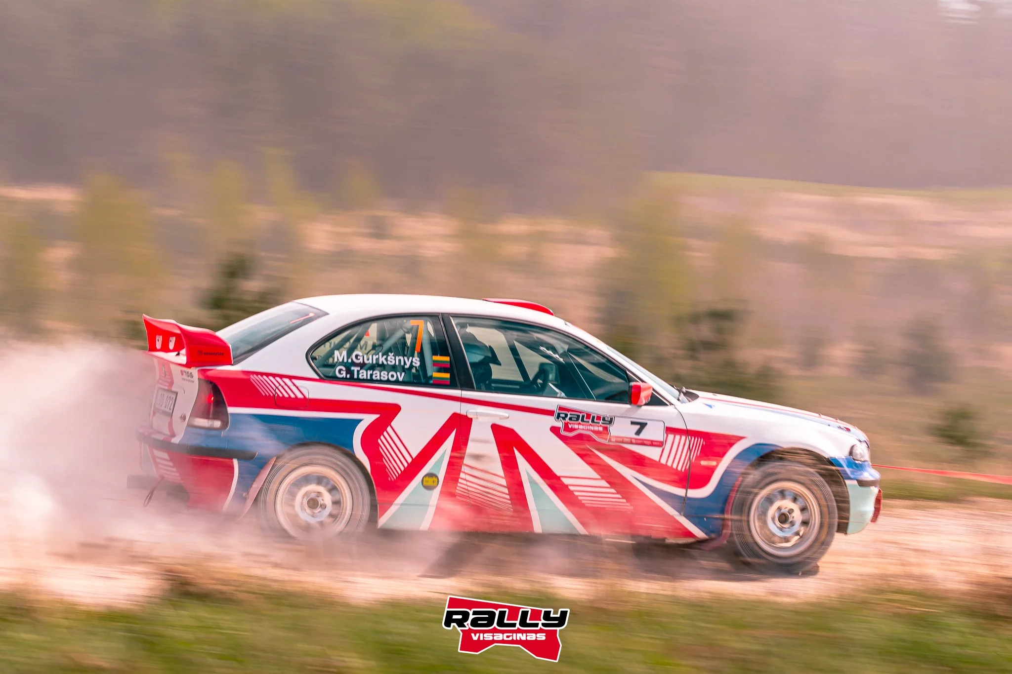 A rally car with a red, white, and blue Union Jack design speeding across a dirt road with dust behind it, driven by M. Gurkšņs and G. Tarasov, number 7, racing through a rural landscape.