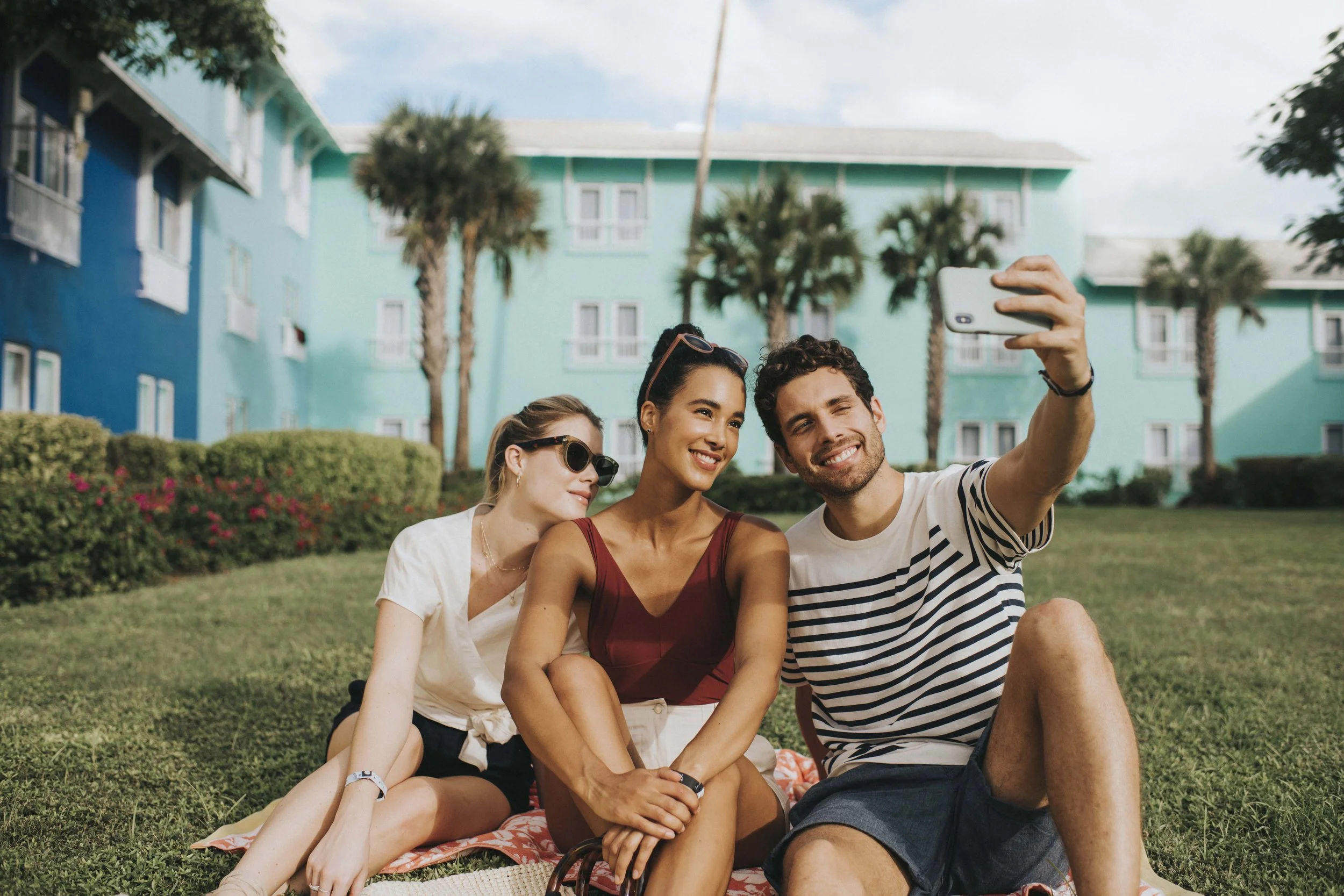 Three models in casual summer outfits taking a selfie on a tropical resort lawn for Club Med, styled by Molly McIntyre. Photographed by Catherine Falls.