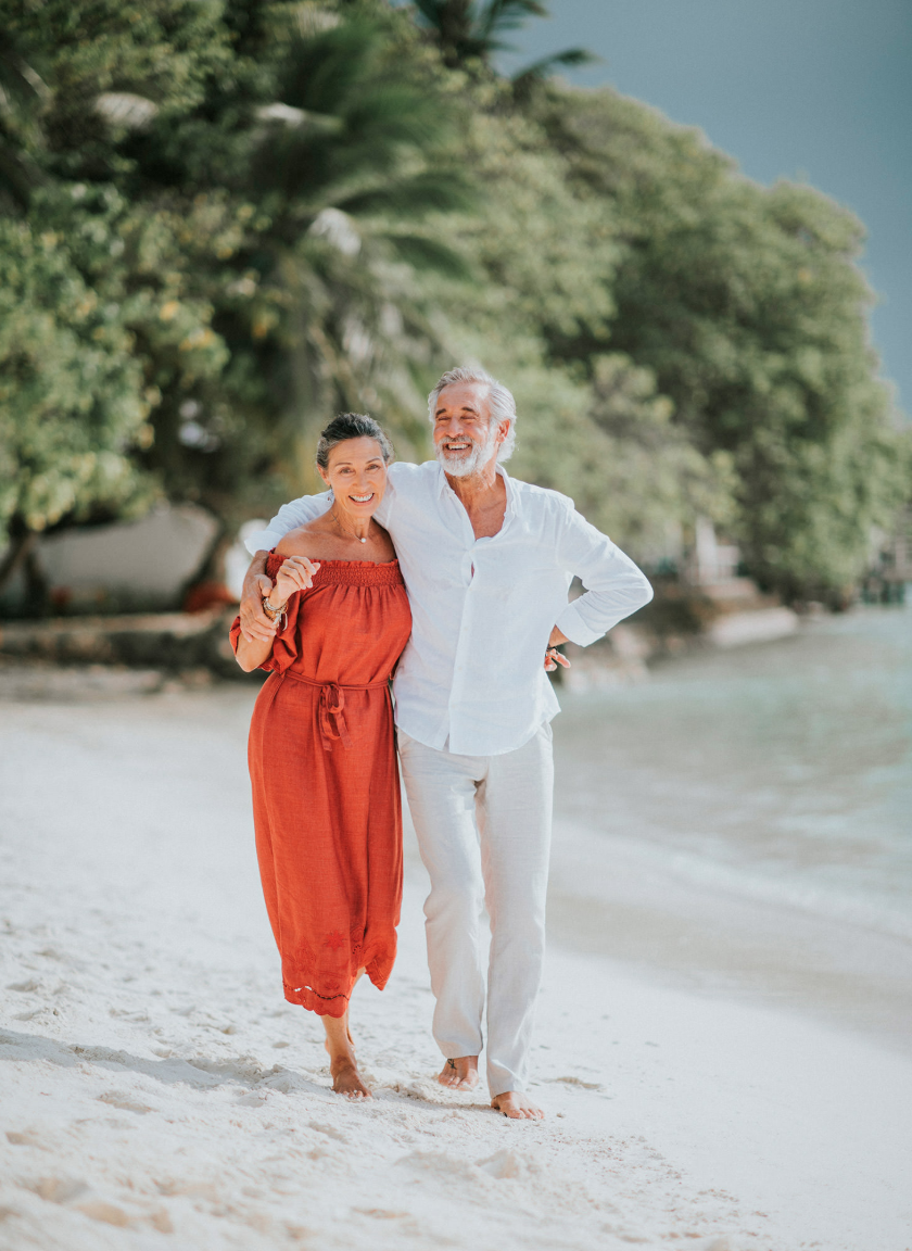 Two models in a rust-orange dress and a white shirt with light trousers walking barefoot on a beach for Club Med, styled by Molly McIntyre. Photographed by Catherine Falls.
