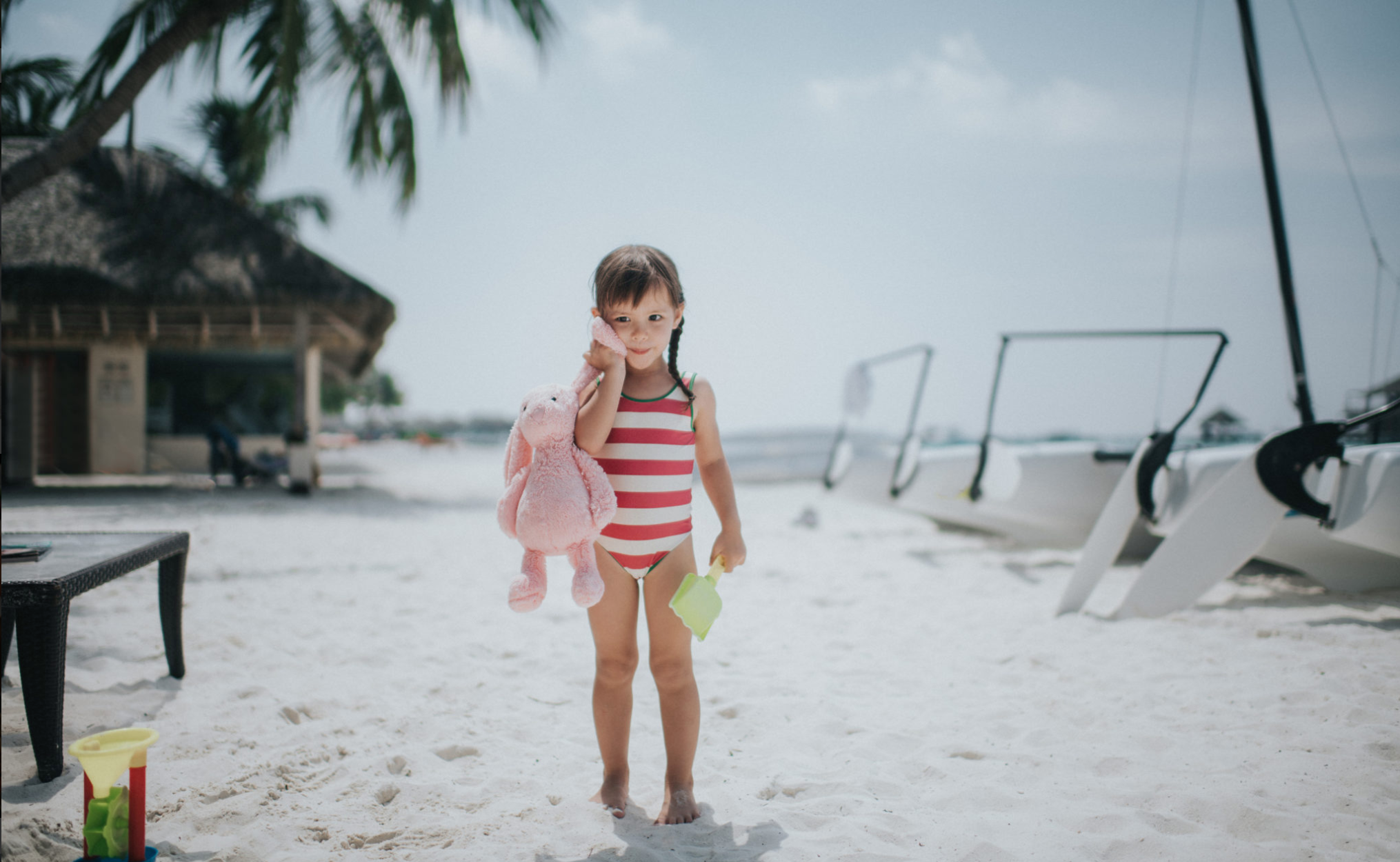Child in a red-and-white striped swimsuit holding a pink toy rabbit and green spade on a beach for Club Med, styled by Molly McIntyre. Photographed by Catherine Falls.