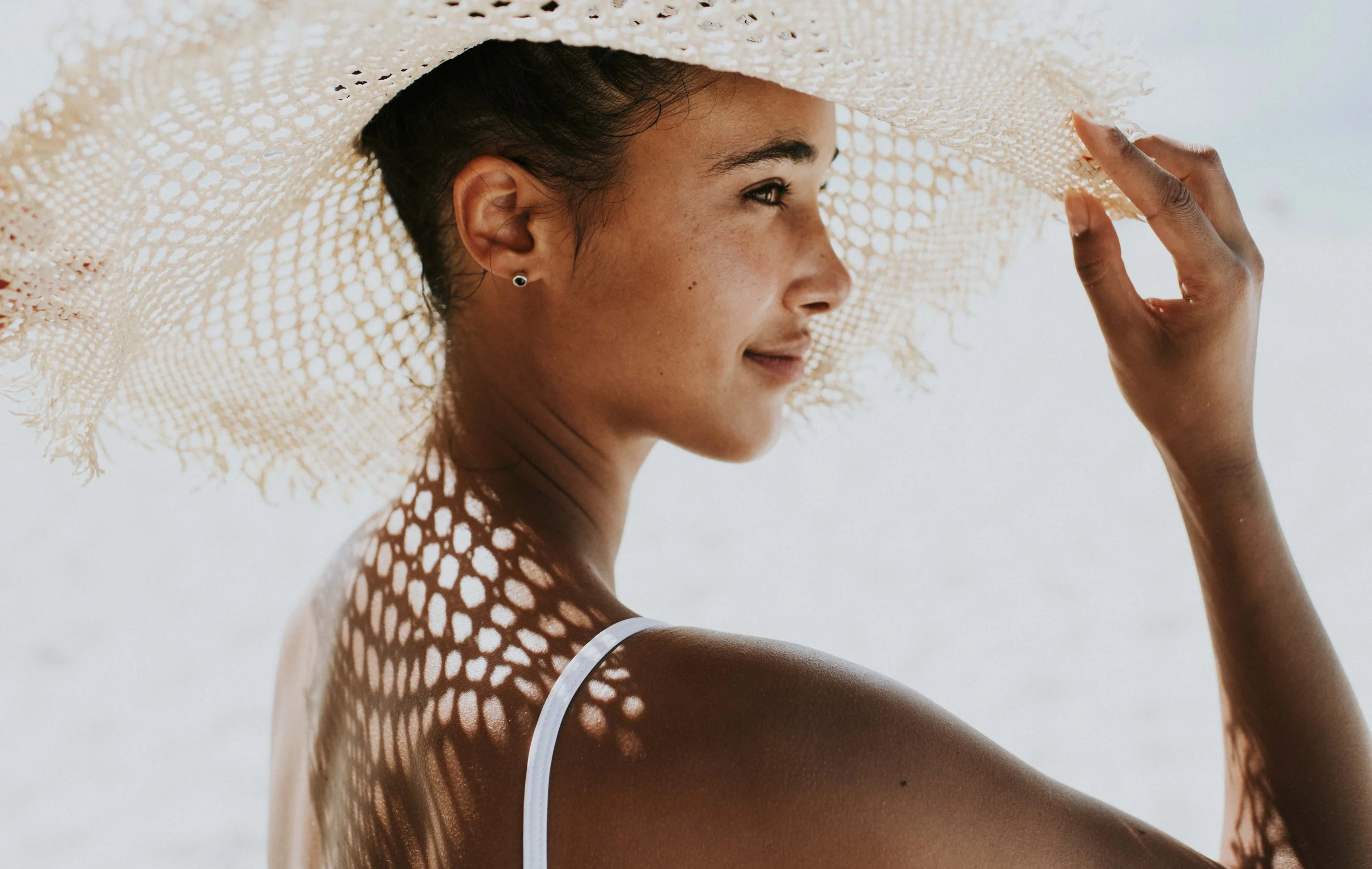 Close-up of a model holding a cream woven sunhat, casting patterned shadows across her shoulder, for Club Med, styled by Molly McIntyre. Photographed by Catherine Falls.