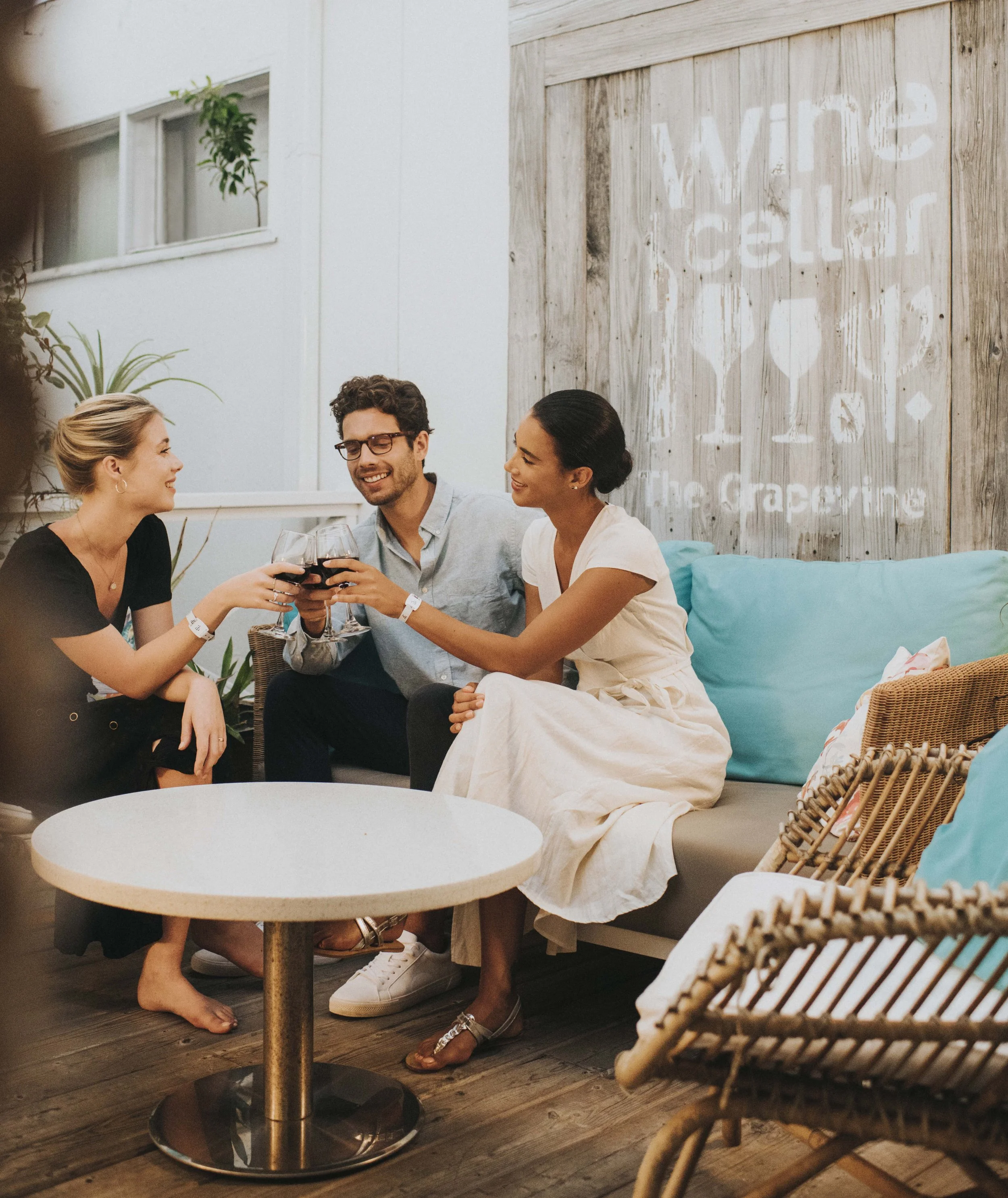 Three models in black and cream dresses and a pale blue shirt sharing a wine toast on a terrace for Club Med, styled by Molly McIntyre. Photographed by Catherine Falls.