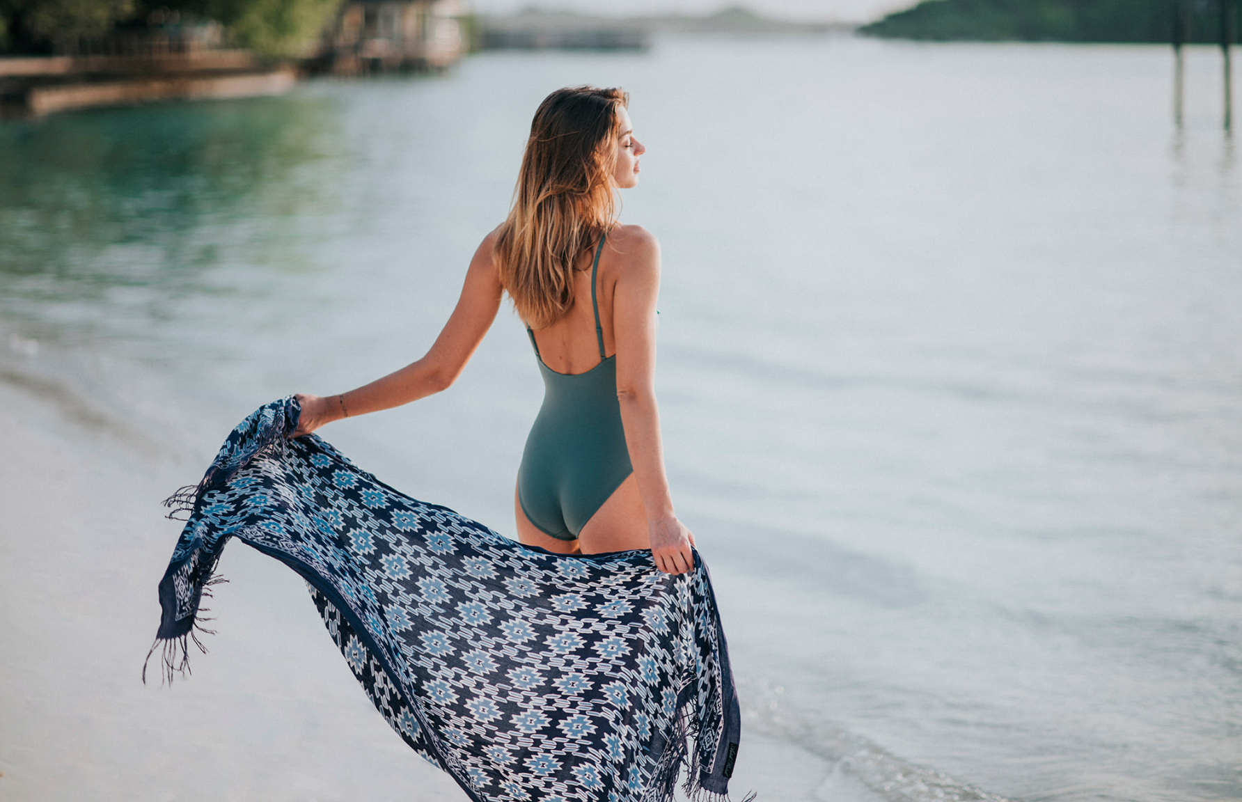 Model in a green swimsuit holding a blue patterned sarong beside calm tropical waters for Club Med, styled by Molly McIntyre.