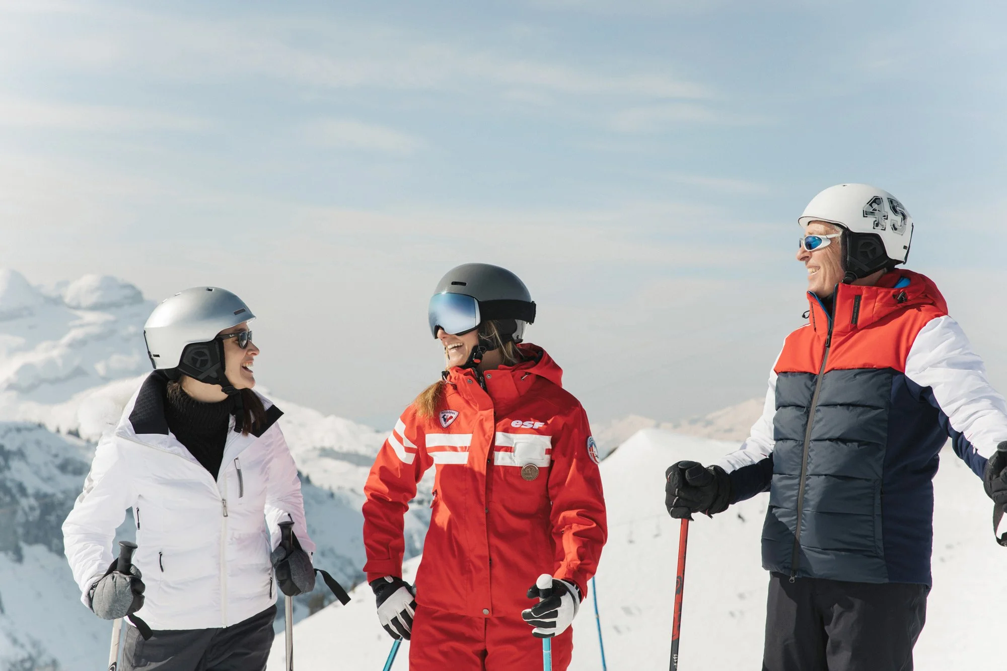 Three skiers in white, red and navy ski outfits smiling together against snowy mountains for Club Med, styled by Molly McIntyre. Photographed by Jules Davies.