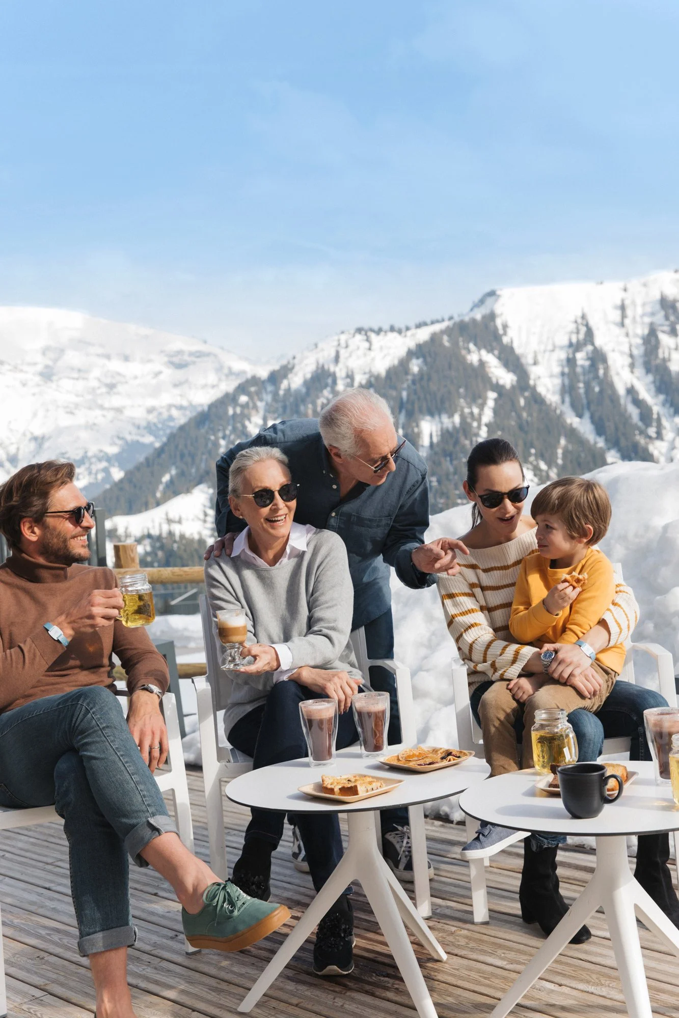 Adults and a child in winter knitwear chatting with a resort staff member on a snowy mountain terrace for Club Med, styled by Molly McIntyre. Photographed by Jules Davies.