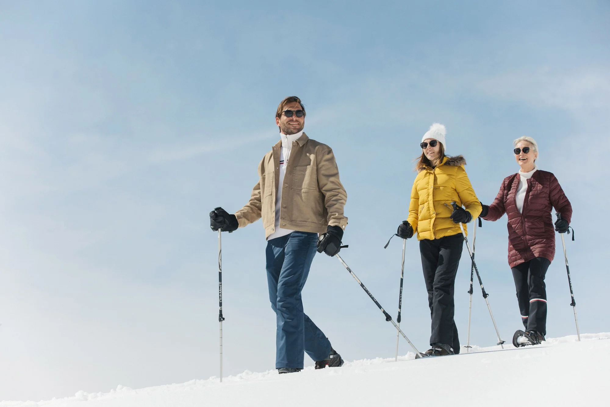 Three adults in beige, yellow and burgundy winter jackets snowshoeing with walking poles for Club Med, styled by Molly McIntyre. Photographed by Jules Davies.
