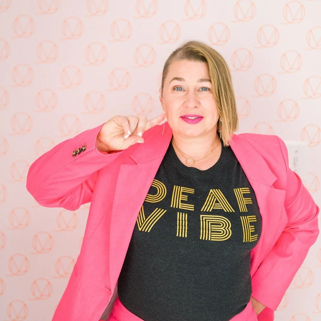Erin is wearing a hot pink suit with a black tee that has "deaf vibe" in gold lettering - art deco style. she is signing the word deaf