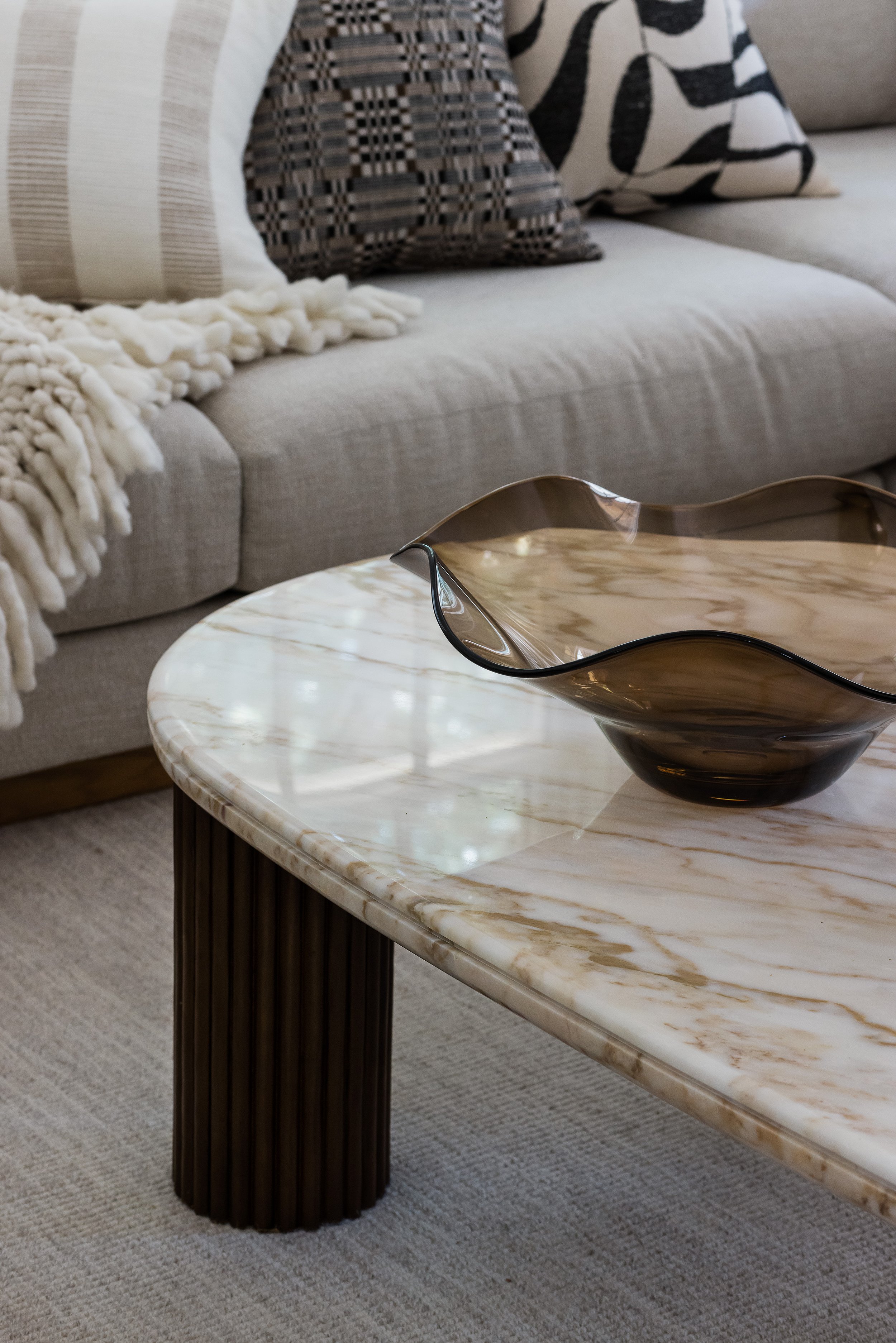 Close-up of a curved travertine coffee table with fluted wood legs and a smoked glass wavy bowl.
