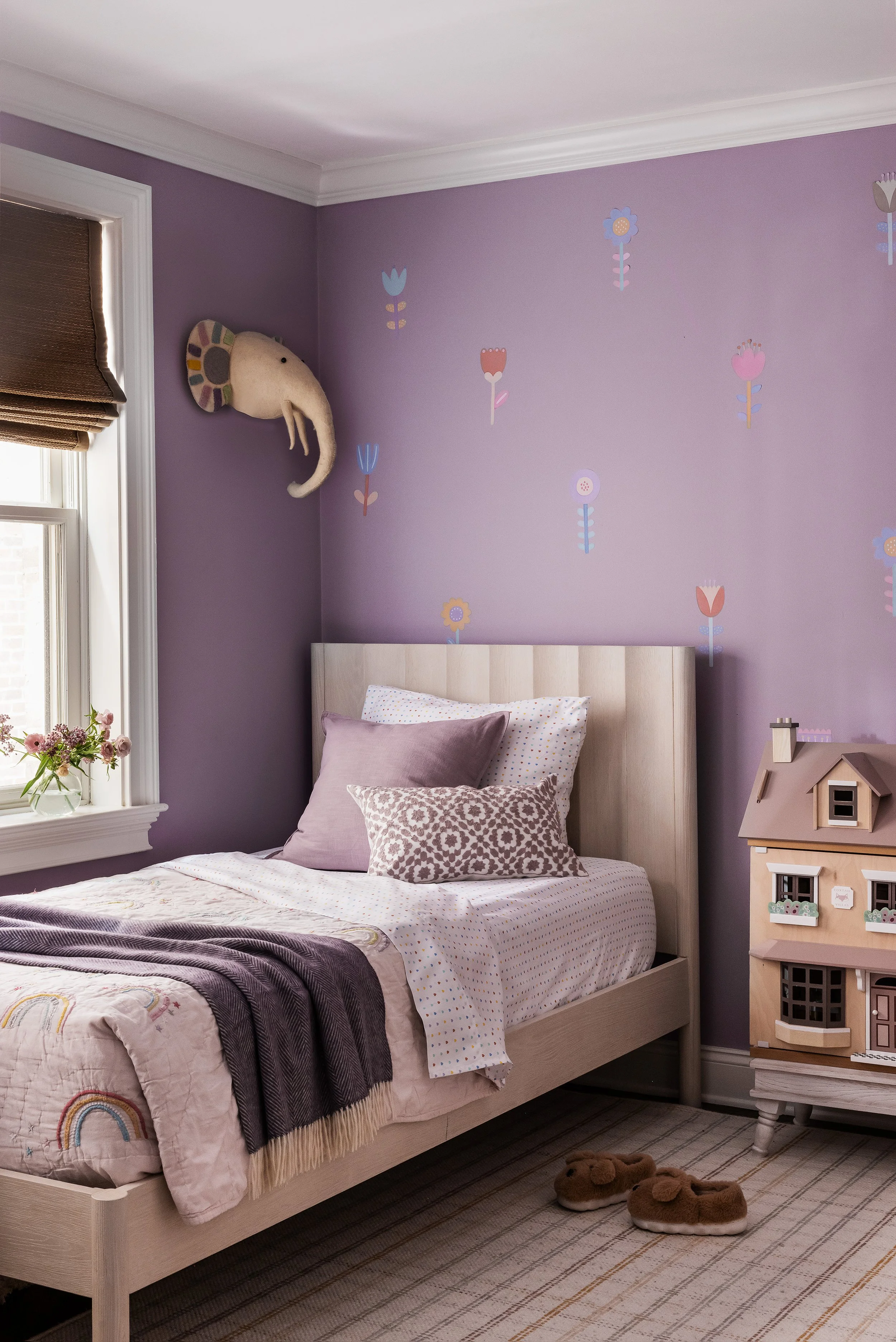 Child's bedroom with lavender walls and floral decals, a light wood upholstered bed, rainbow quilt, and a dollhouse beside the window.