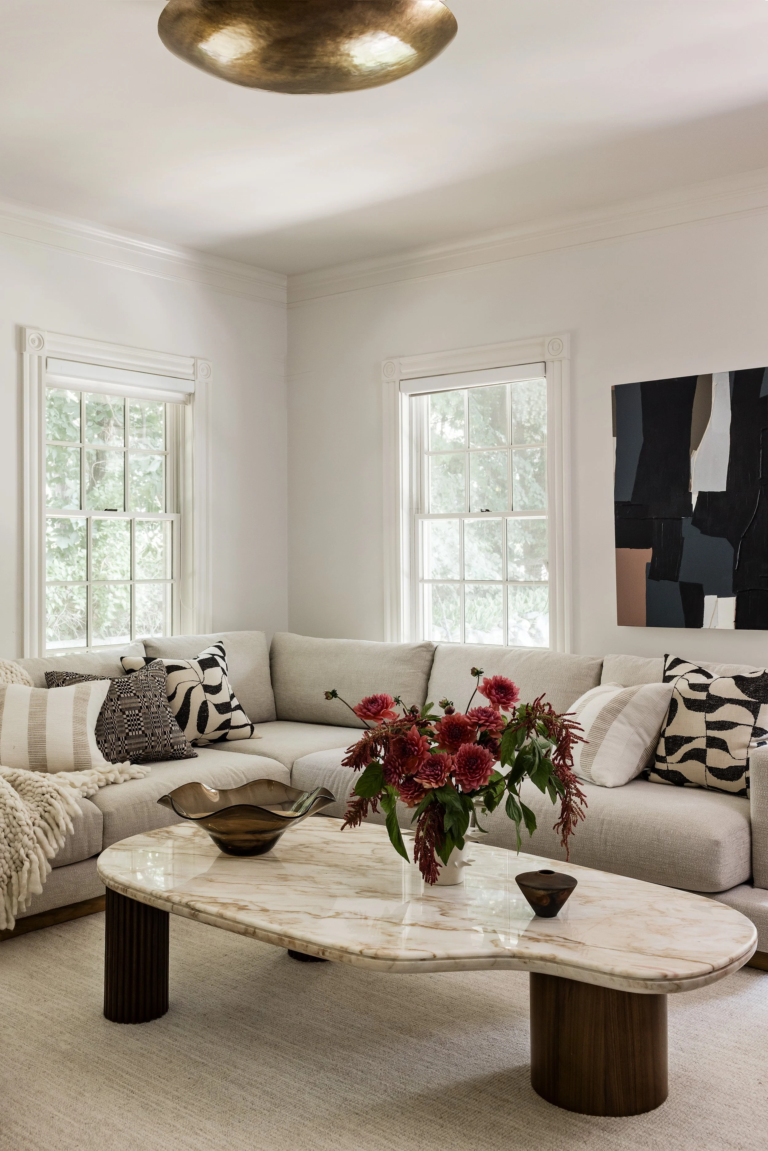Living room with a light gray sectional, black and white graphic throw pillows, a curved travertine coffee table, and a dark abstract painting.