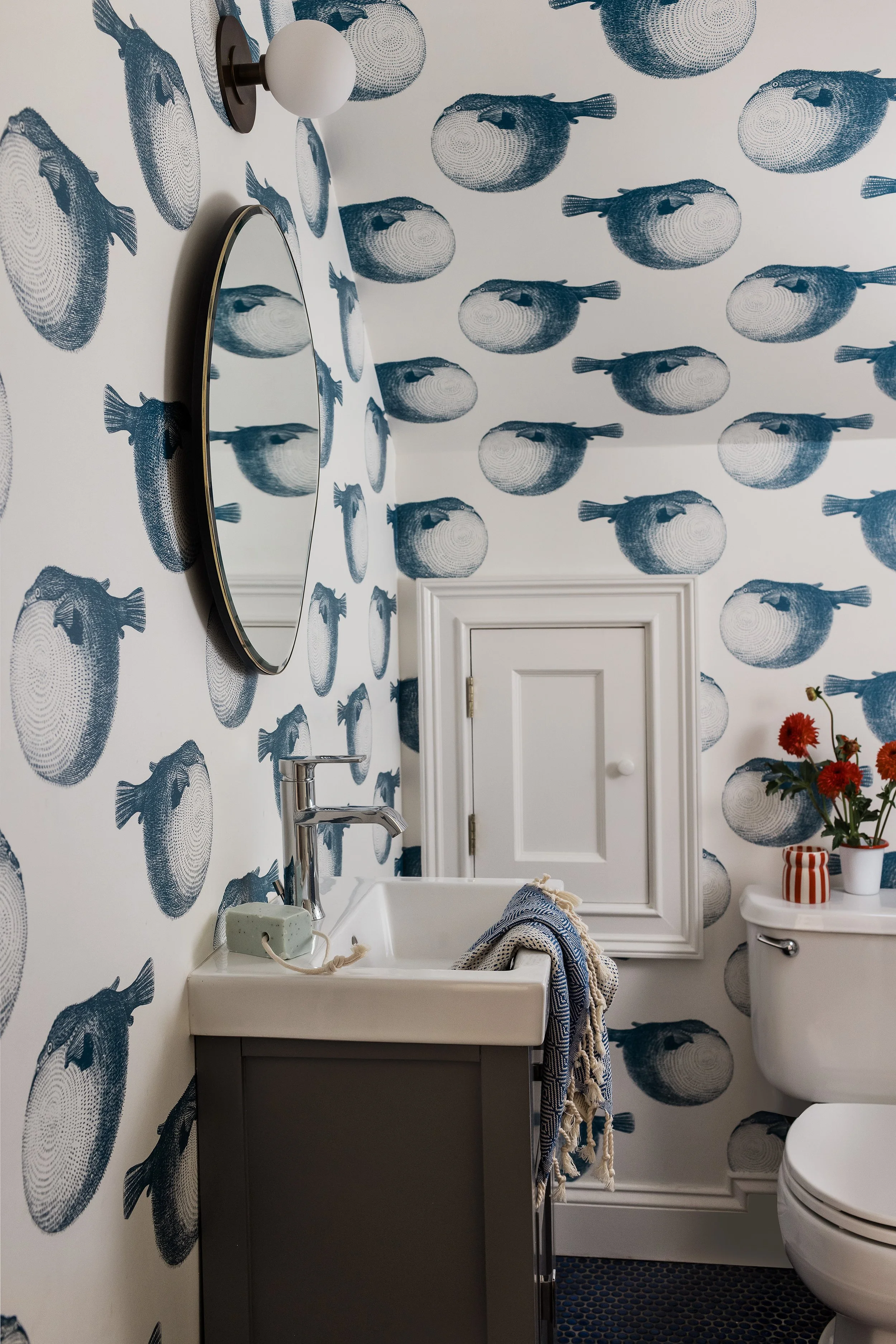Powder room fully wrapped in blue pufferfish print wallpaper, walls to ceiling, with a round brass-framed mirror and a small white vanity.