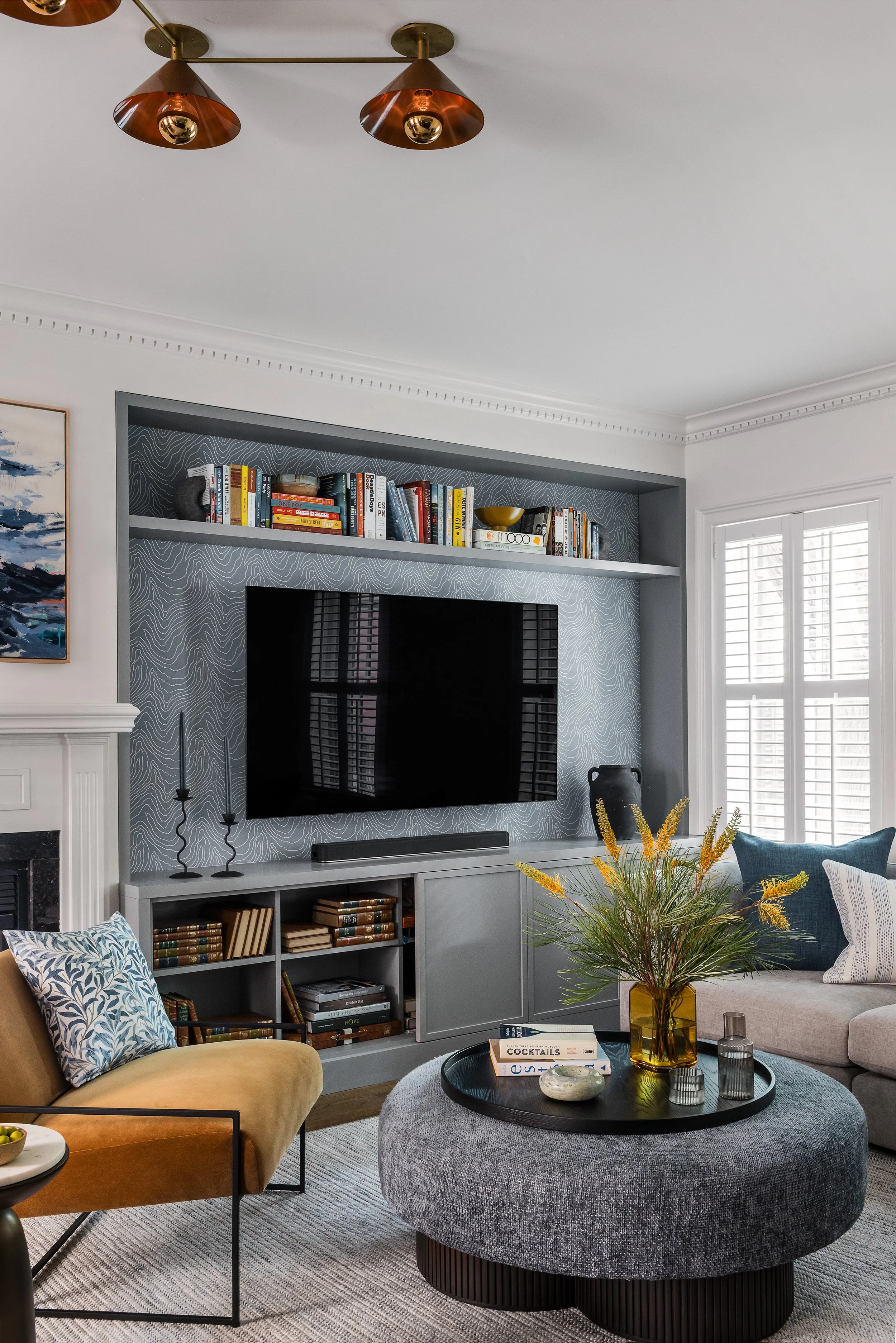 Vertical detail of a gray media console and wallpapered TV alcove with black candlesticks, a dark ceramic urn, and open bookshelves below.
