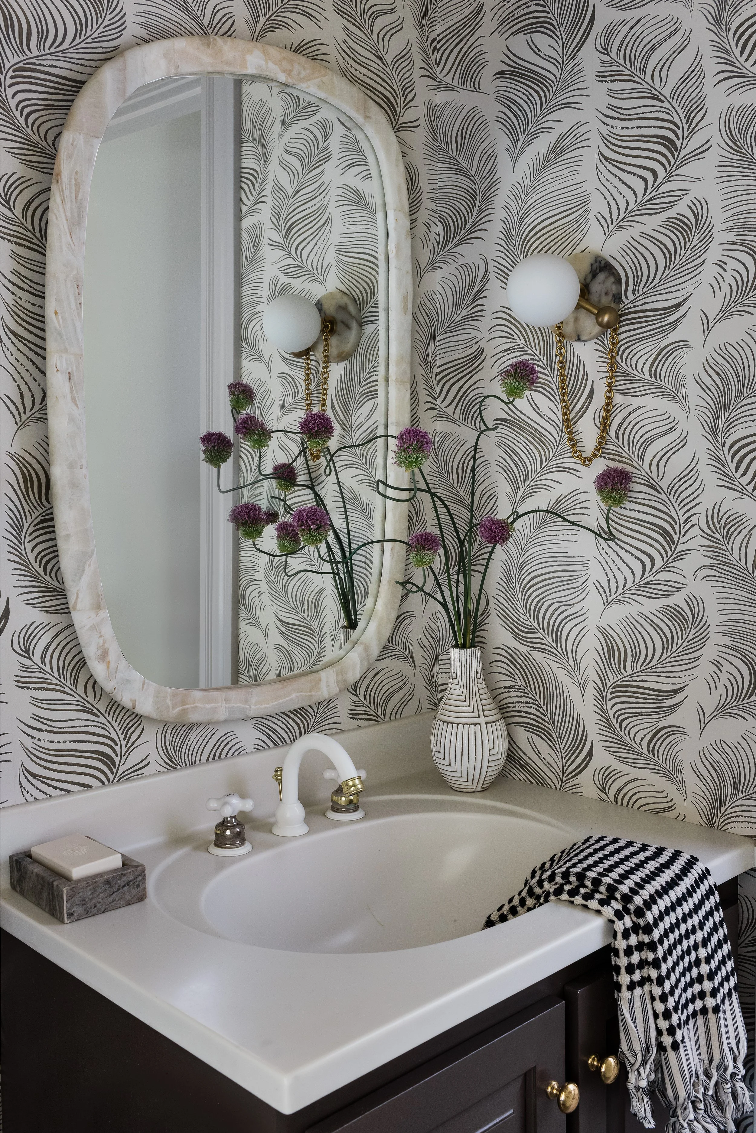 Powder room with black and white feather print wallpaper, an oval stone-framed mirror, marble sconces, and a textured ceramic vase.