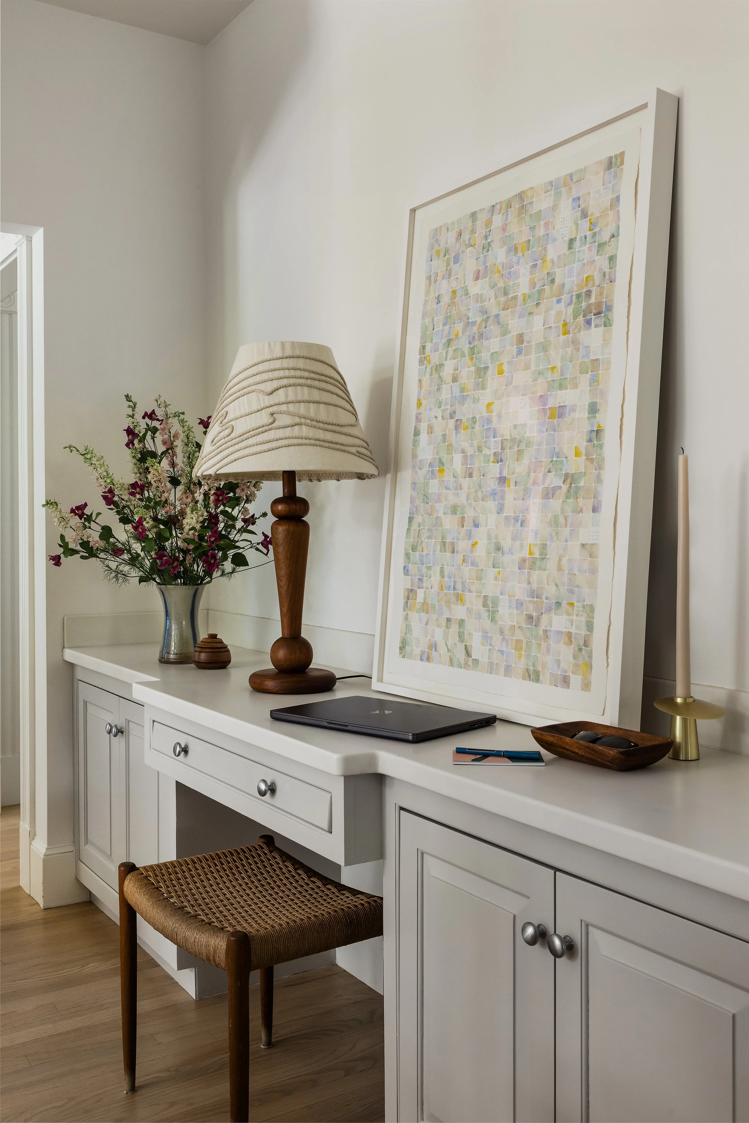 Small home office desk with a turned wood lamp, woven stool, and a large abstract watercolor collage leaning against the wall.