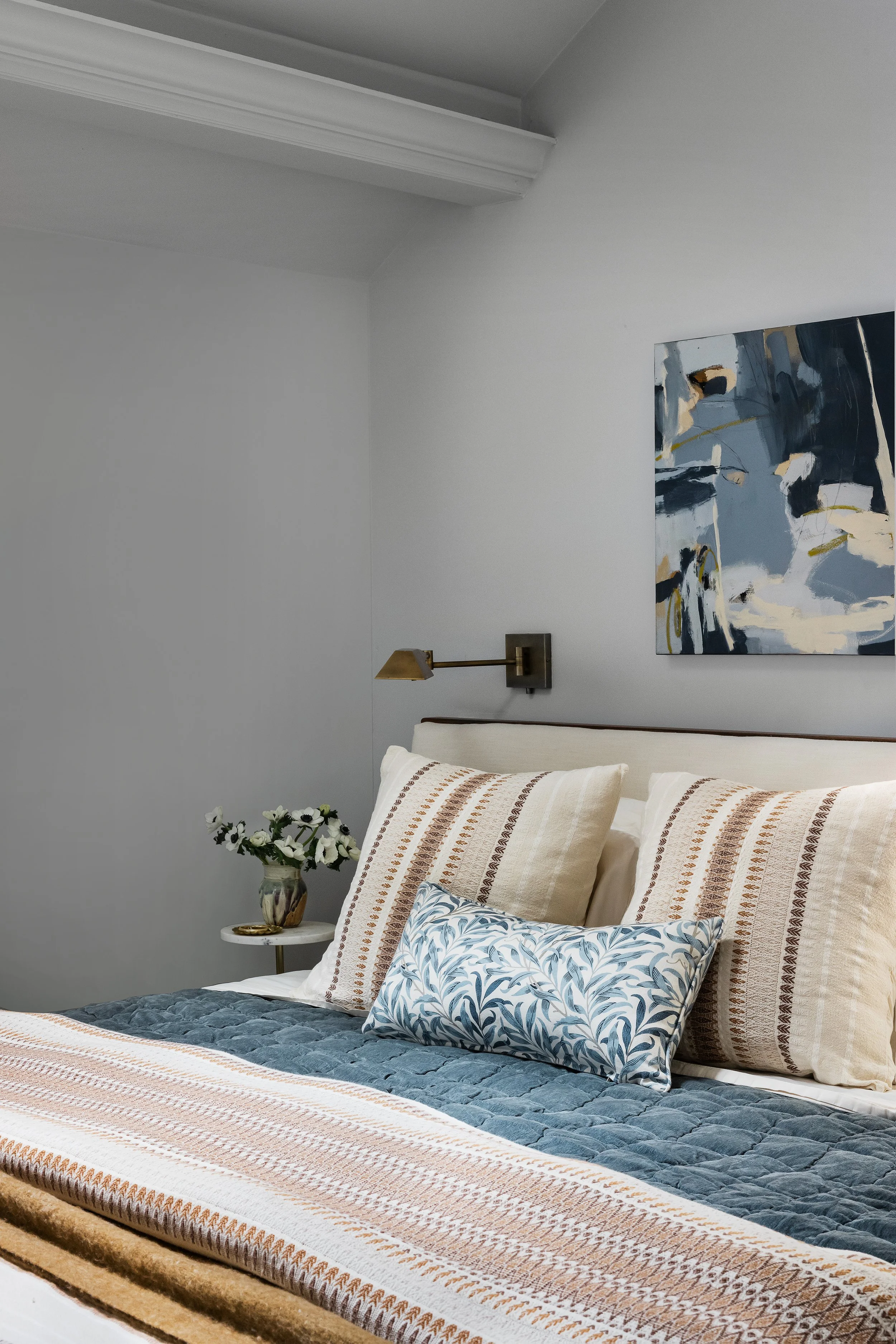 Primary bedroom detail with an upholstered headboard, blue quilted coverlet, striped and floral pillows, and an abstract painting above a brass wall sconce.