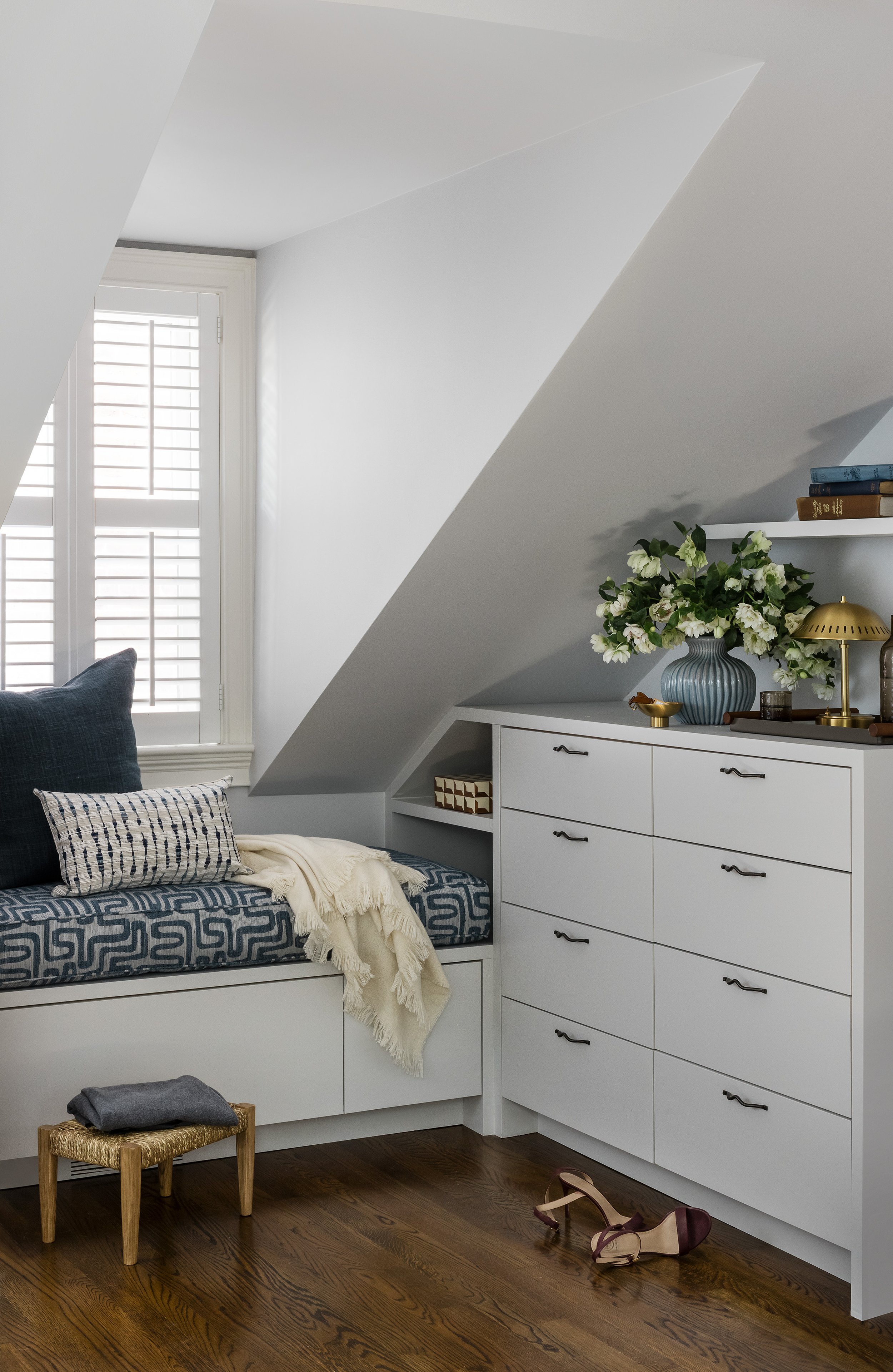Built-in window seat and dresser tucked under a dormer ceiling, with a blue patterned cushion, brass lamp, and fresh flowers on the dresser top.