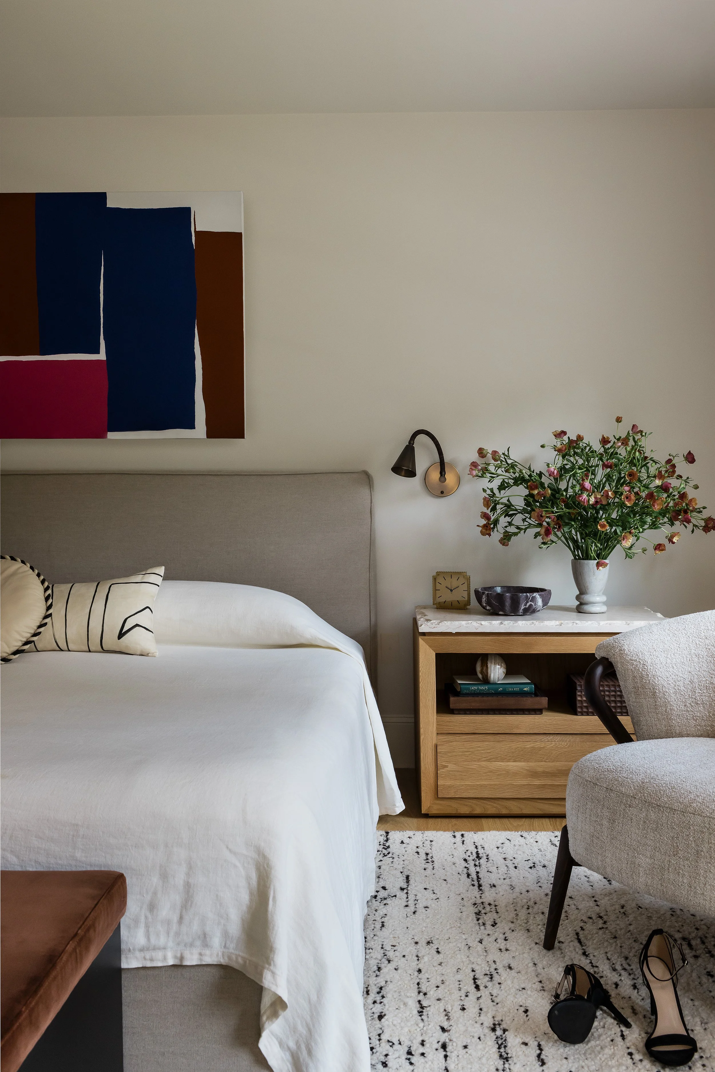 Wood nightstand with a marble top, ceramic bowl, brass clock, and a vase of pink ranunculus beside a linen bed.