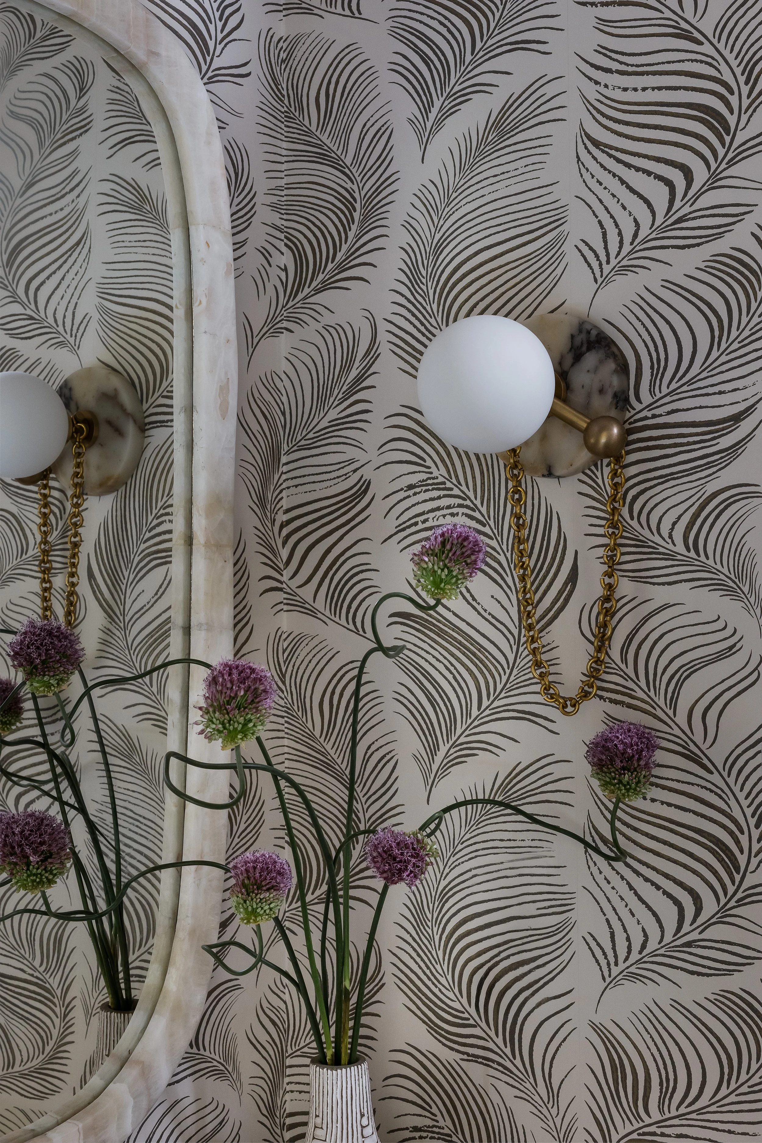 Close-up of a marble wall sconce with a brass chain and globe light fixture against feather print wallpaper, with purple allium in a textured vase.