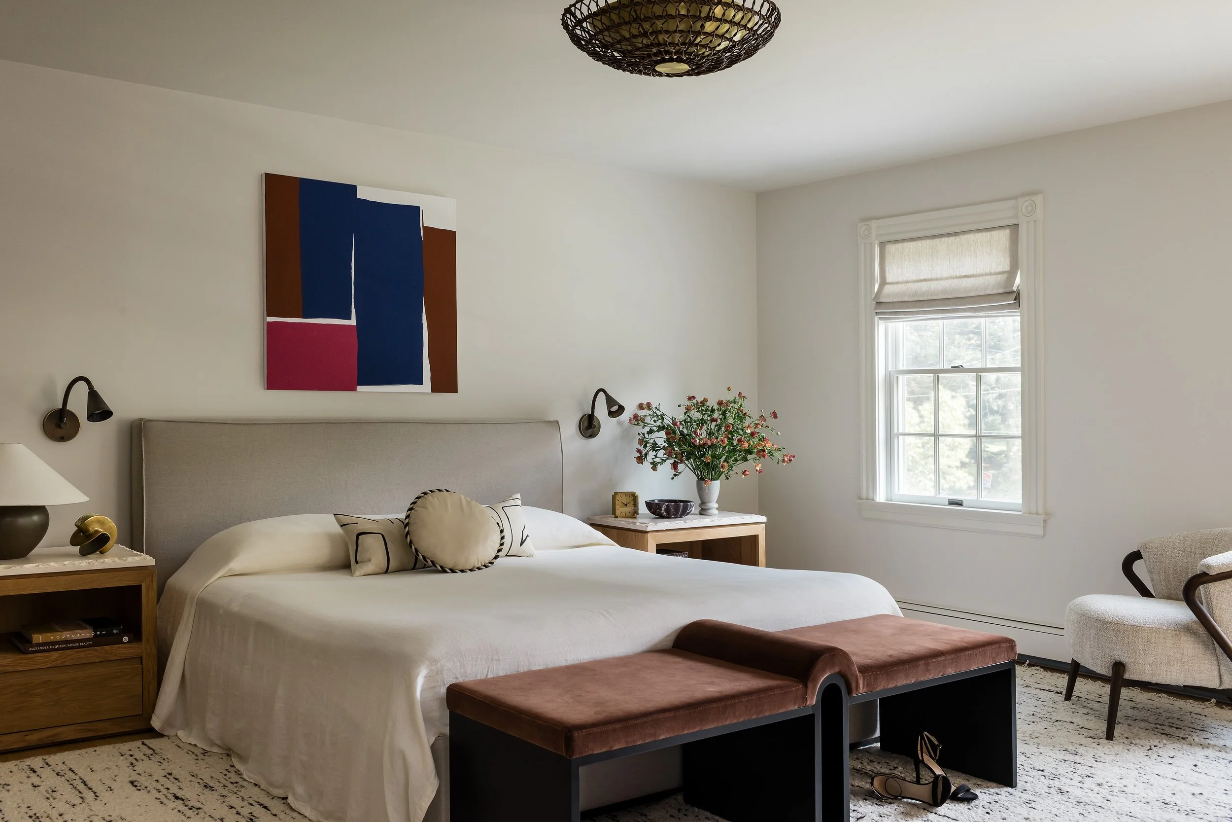 Primary bedroom with a linen upholstered bed, brown velvet bench, boucle armchair, and a color block abstract painting in navy, rust, and magenta.