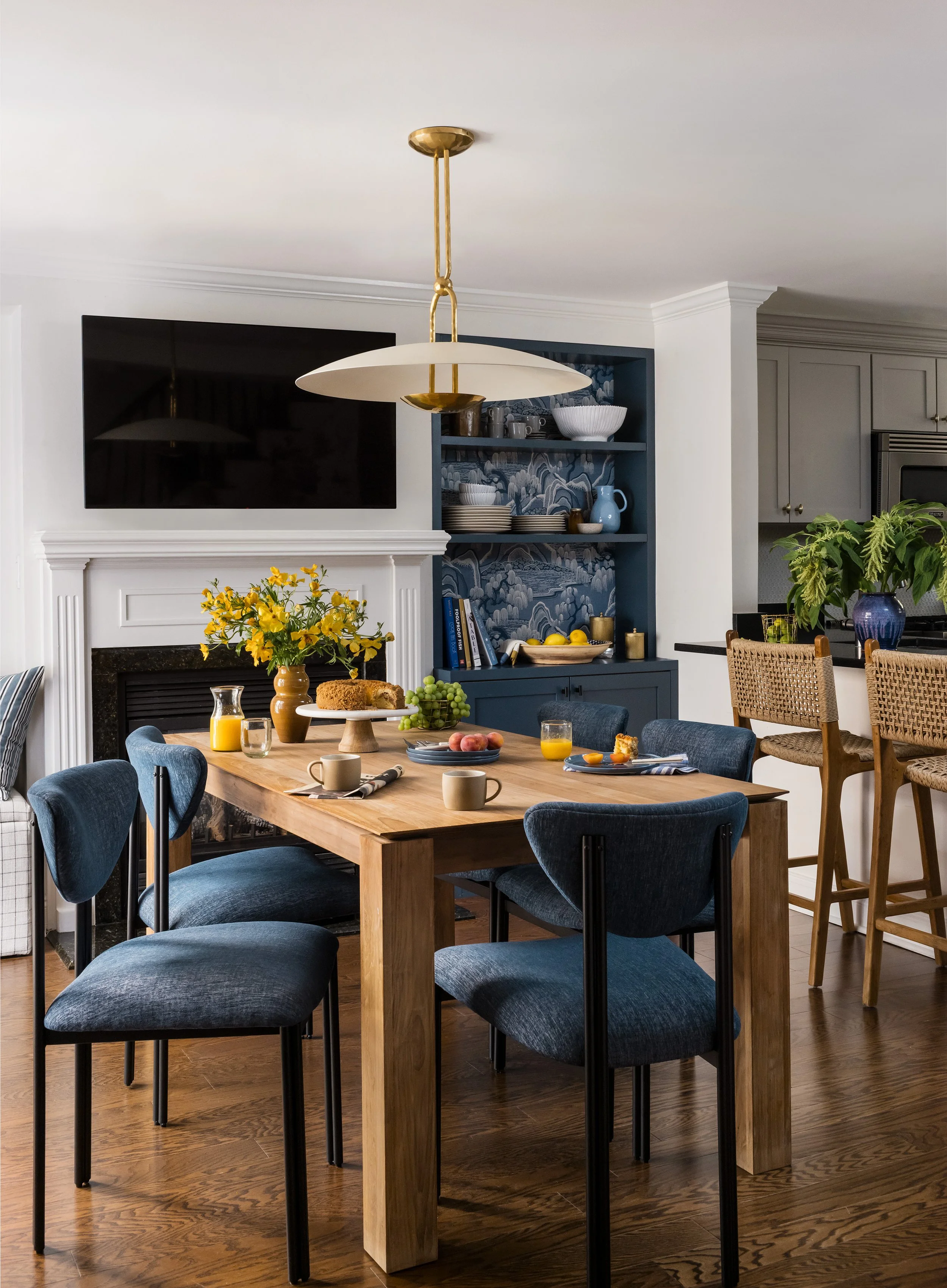 Dining area with a wood table, blue upholstered chairs, a brass dome pendant, and a navy built-in hutch lined with chinoiserie wallpaper, opening to the kitchen.