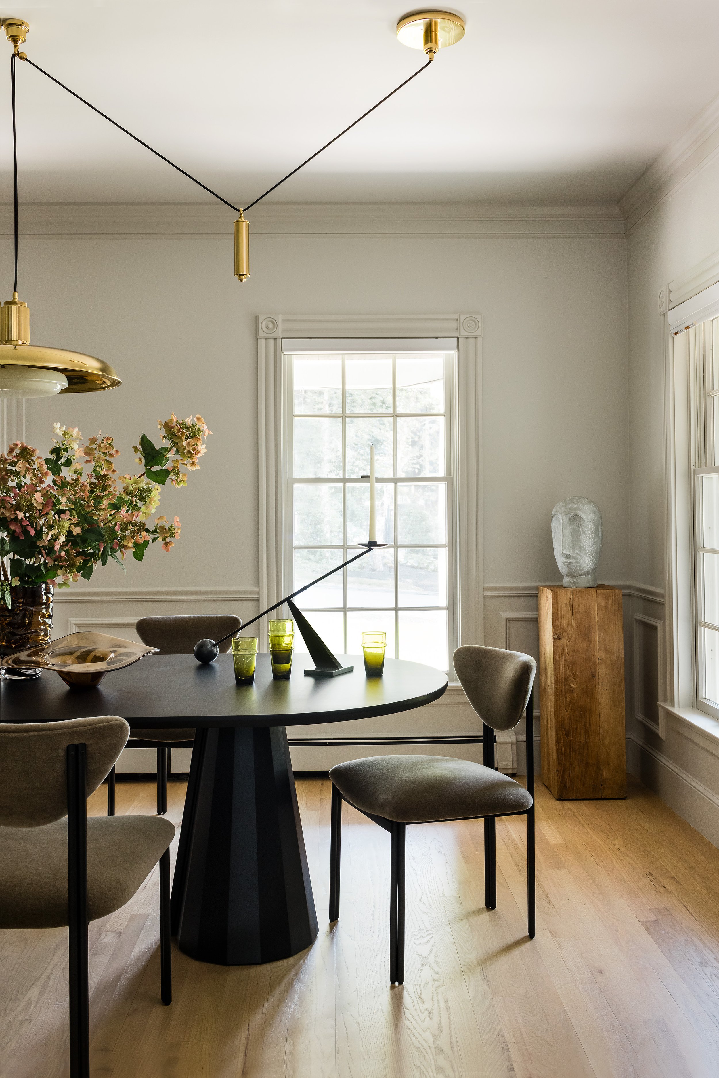Dining room with a black oval table, taupe velvet chairs, a brass sculptural pendant, and a carved wood pedestal with a plaster head sculpture.