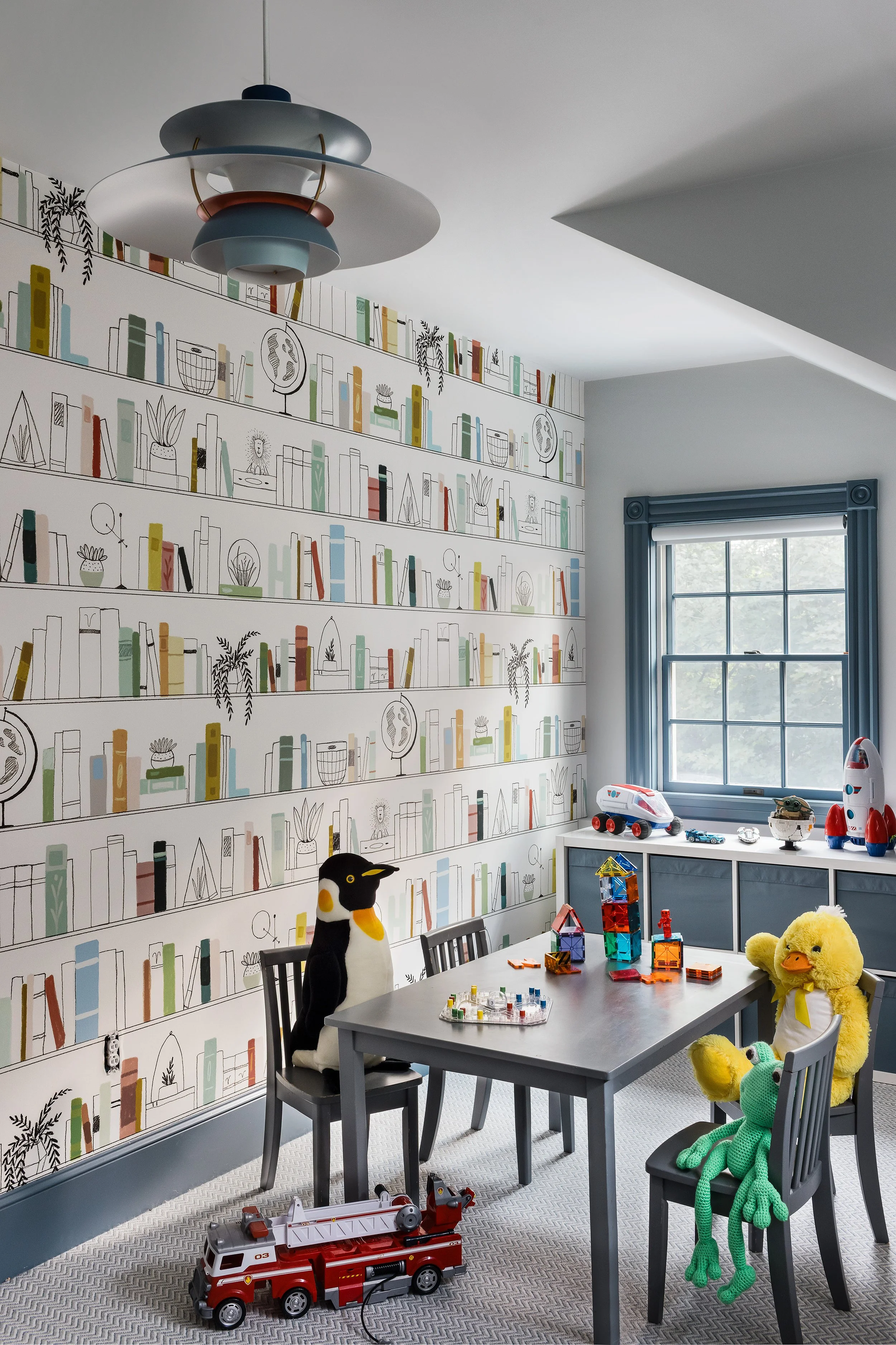Playroom with illustrated bookshelf wallpaper, a gray kids' table and chairs, and a blue tiered pendant light.