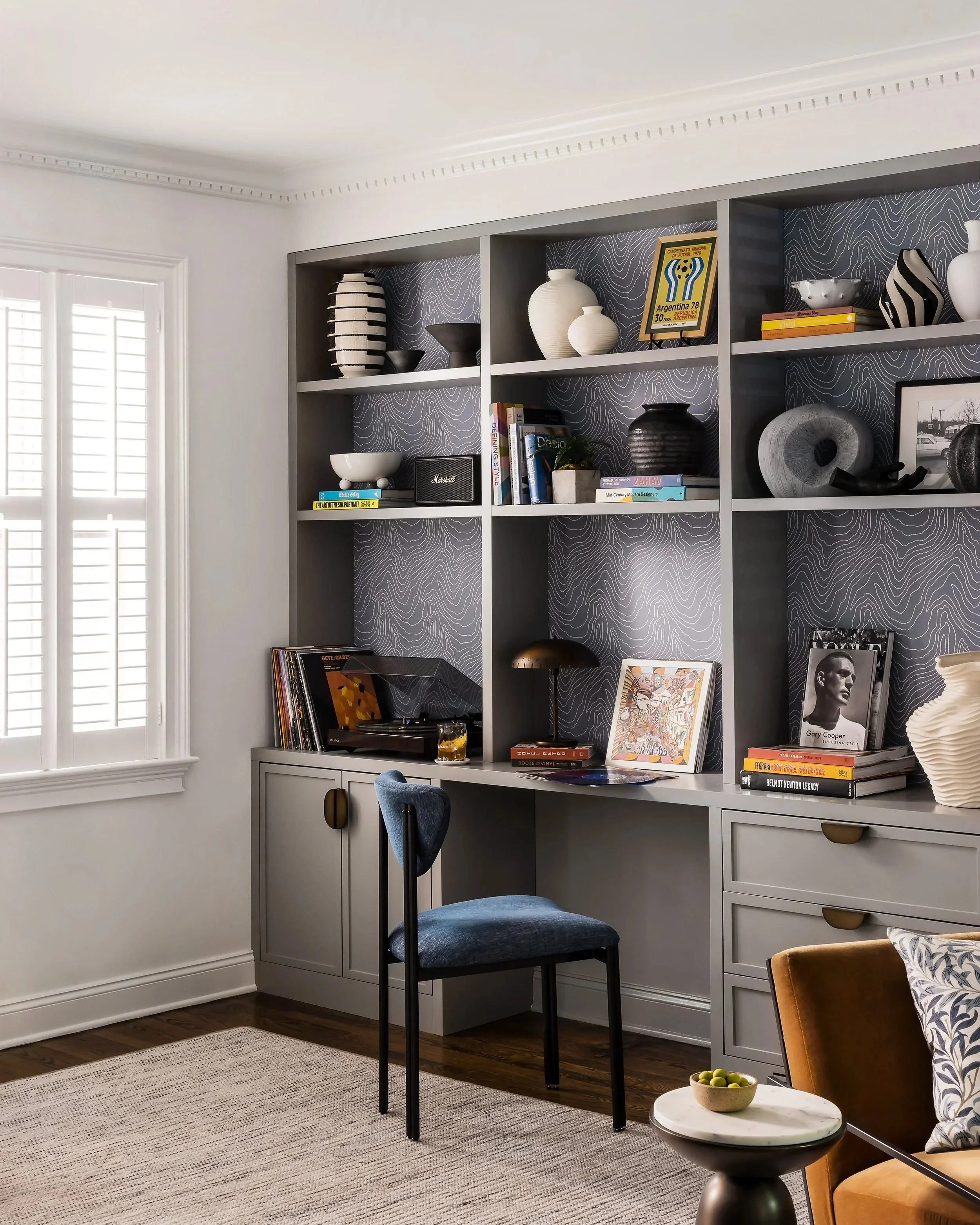 Close-up of a gray built-in desk with a turntable and vinyl storage, brass table lamp, and patterned wallpaper visible behind open shelving.