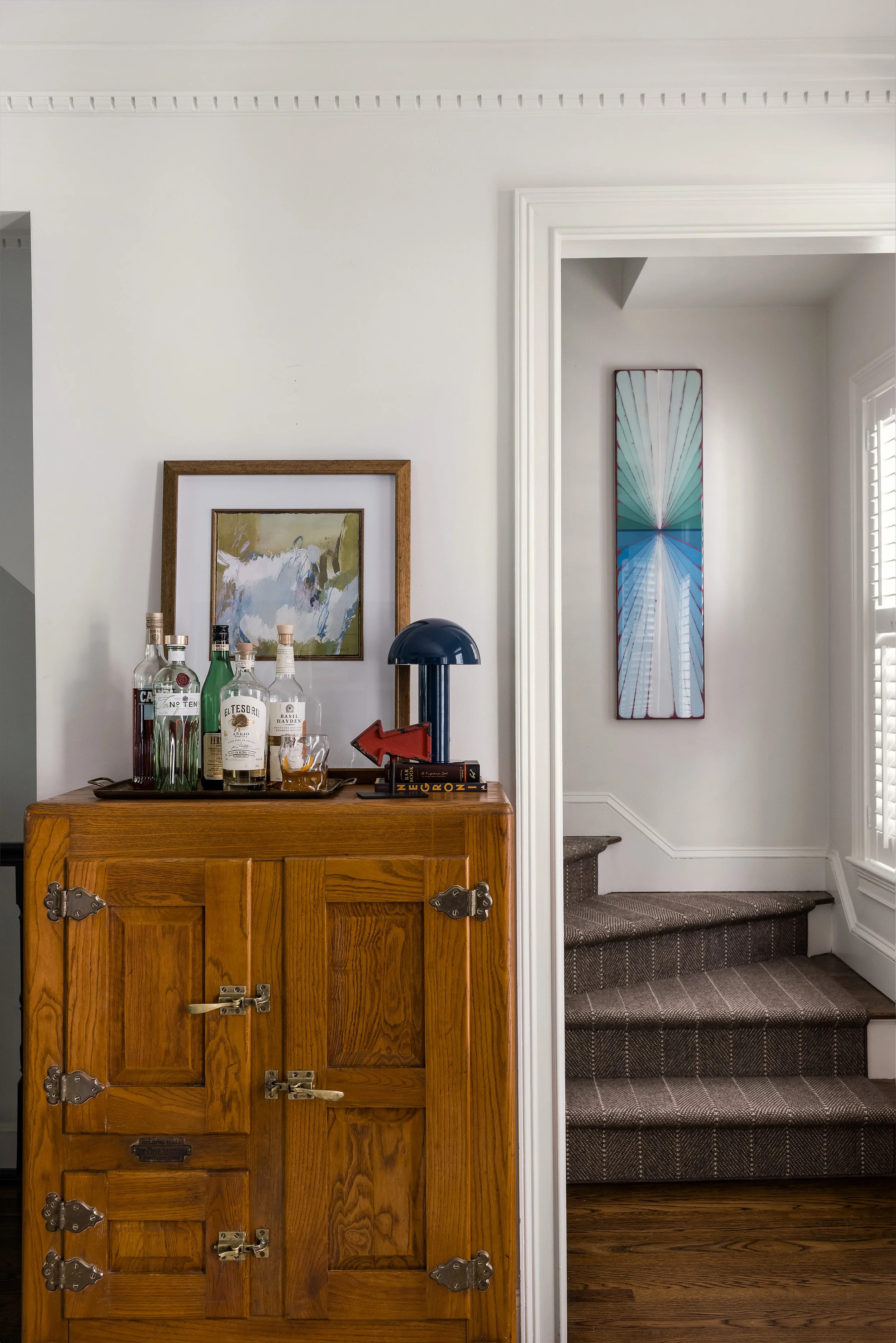 Antique oak icebox repurposed as a bar cabinet, topped with spirits, a cocktail, and a framed abstract painting, with a staircase visible beyond.