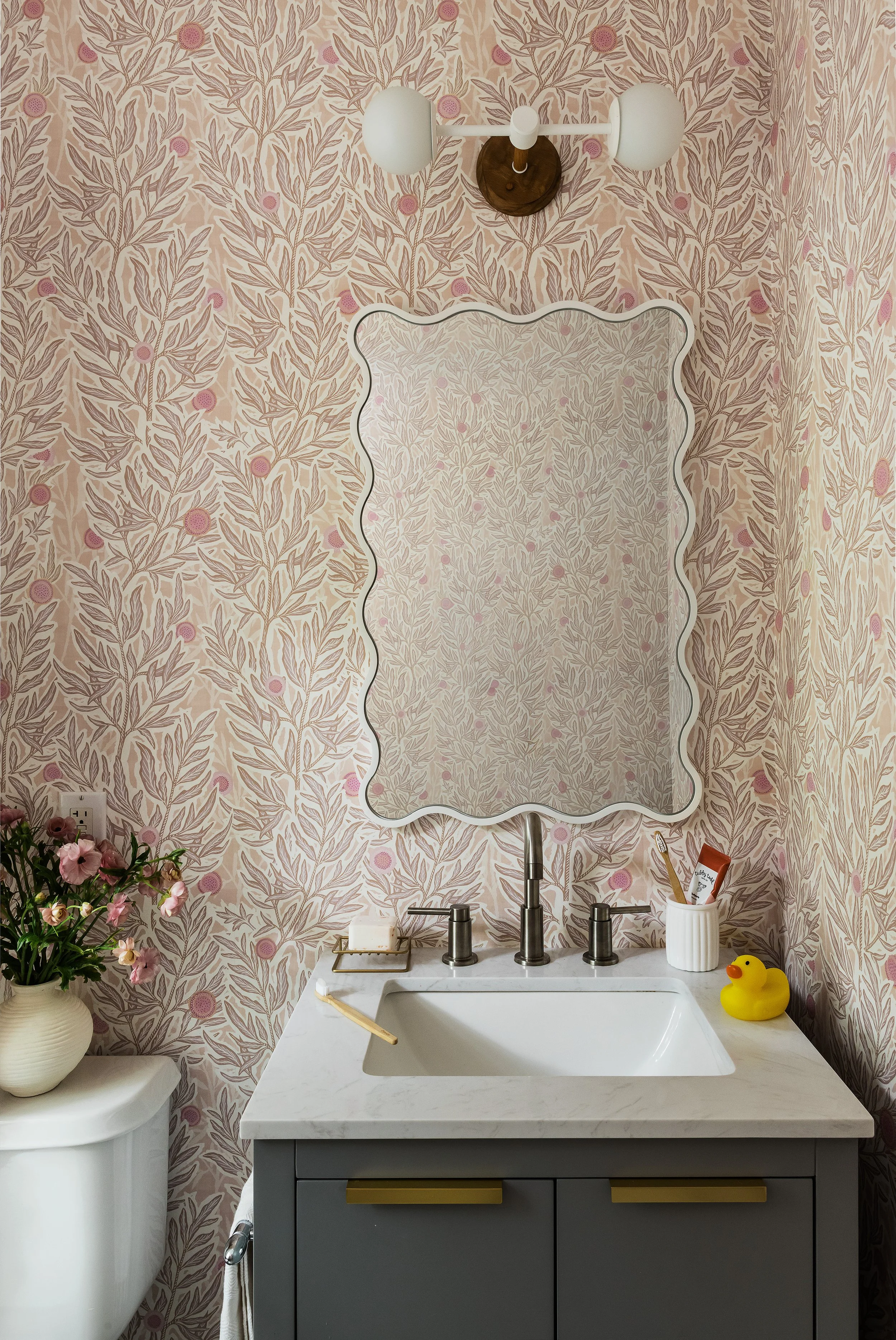 Bathroom with pink and cream leaf print wallpaper, a scalloped white mirror, brass fixtures, and a rubber duck on the vanity.
